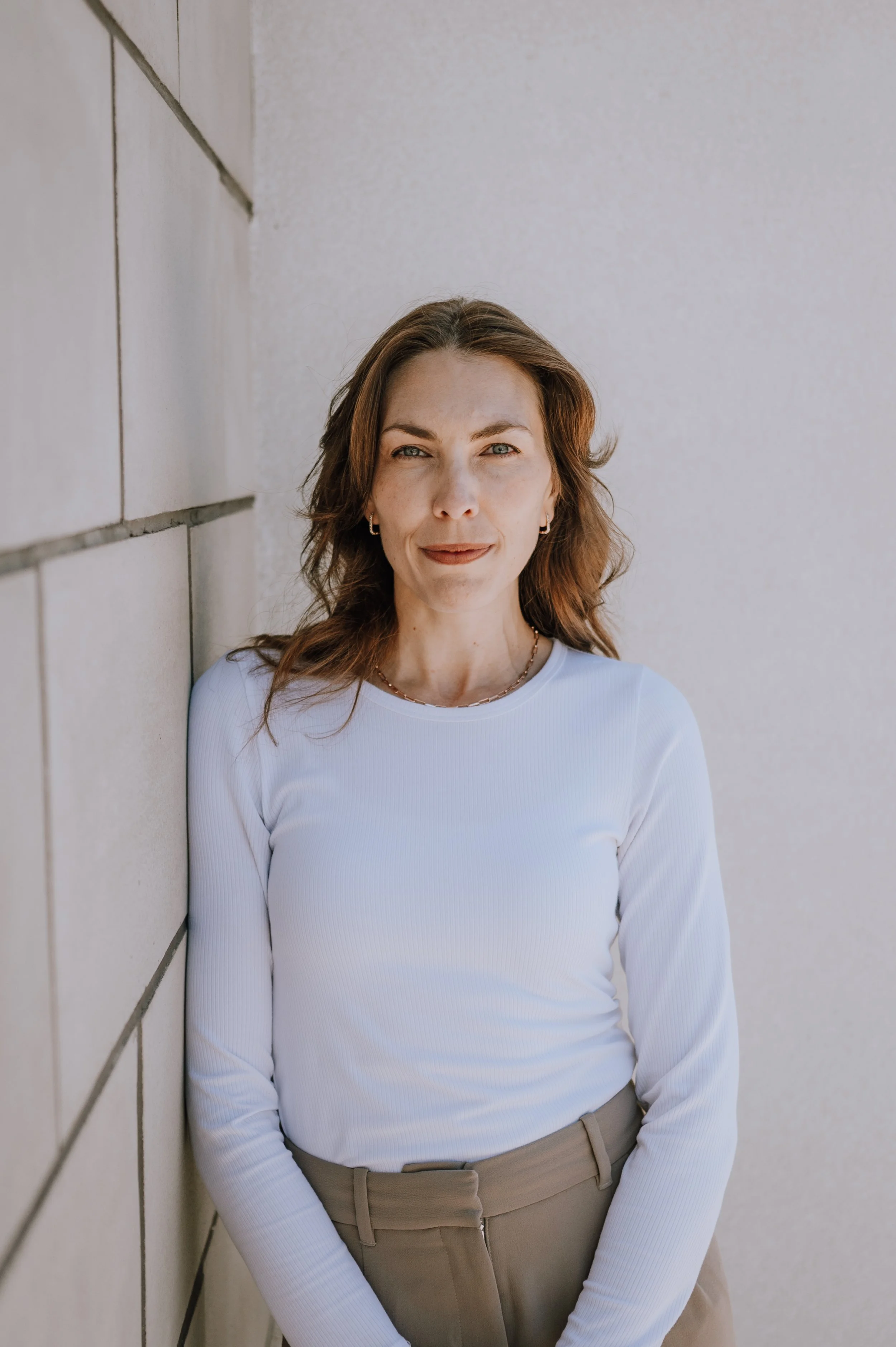 A woman smiling and wearing a white turtleneck and brown pants standing against a plain white background.