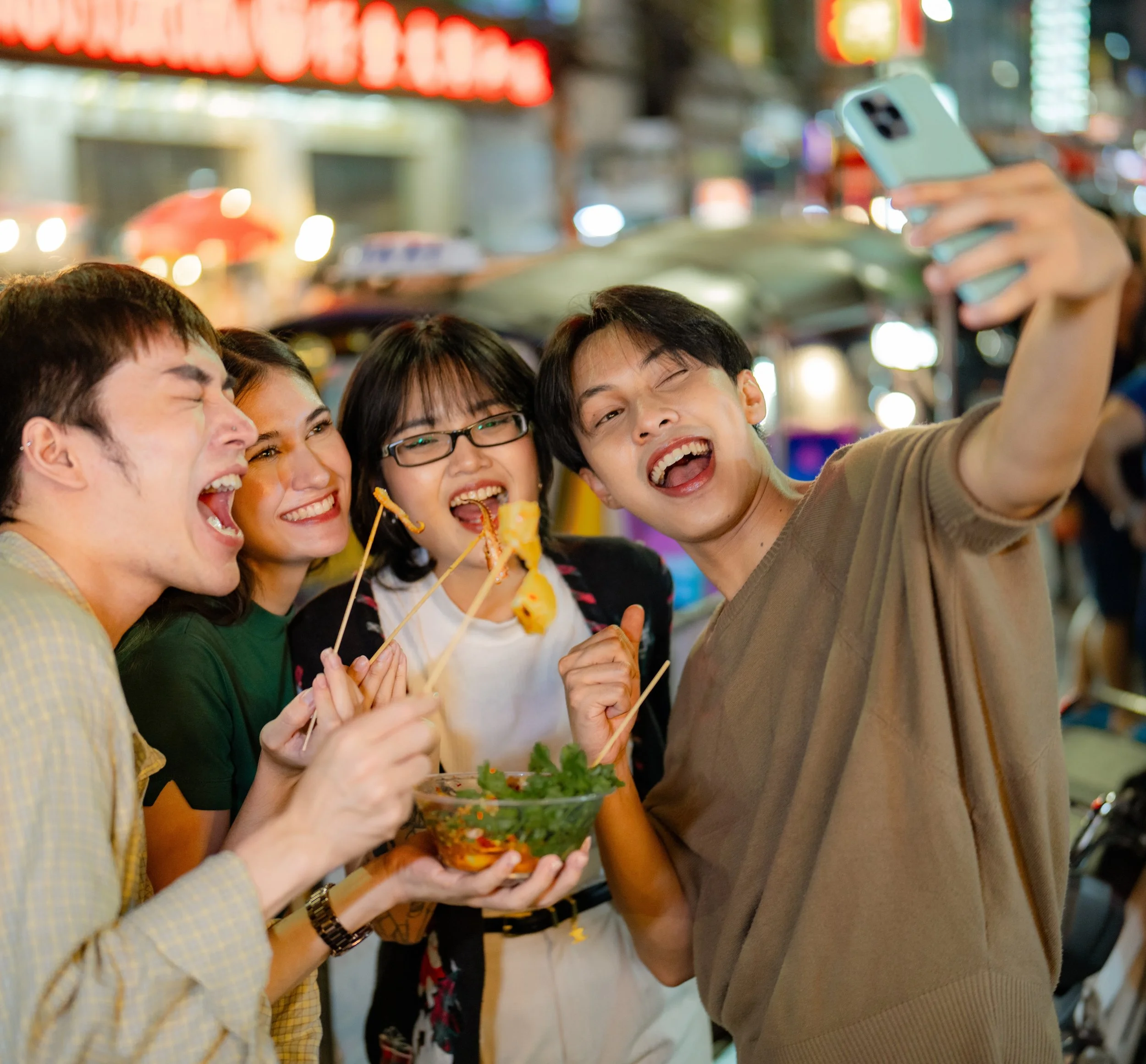 Group of four friends taking a selfie together in a lively street at night, enjoying food and drinks, with colorful lights and signs in the background.