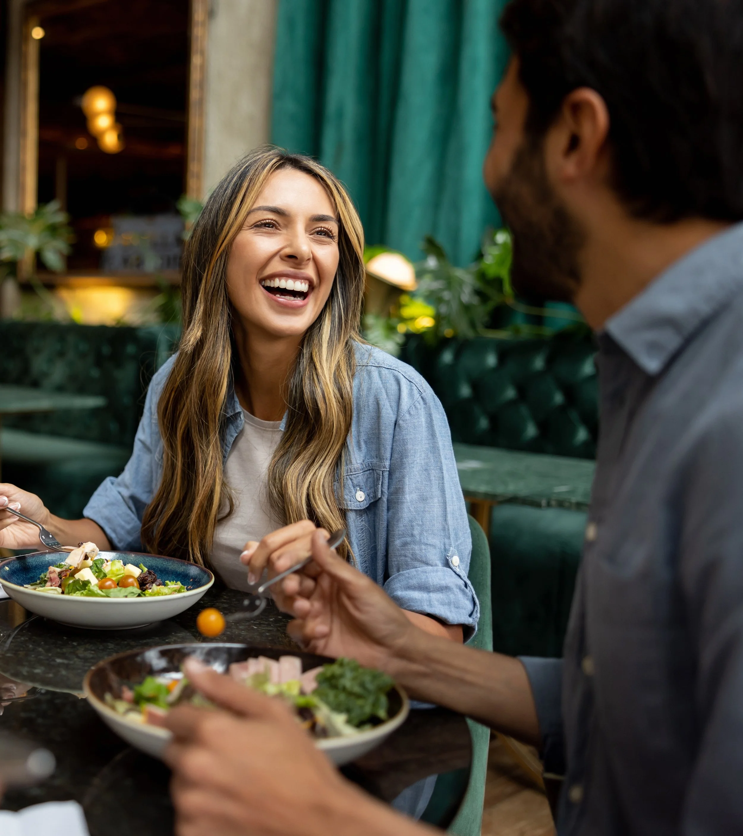 A woman with long blonde hair smiling and laughing while enjoying a salad with a man in a restaurant.