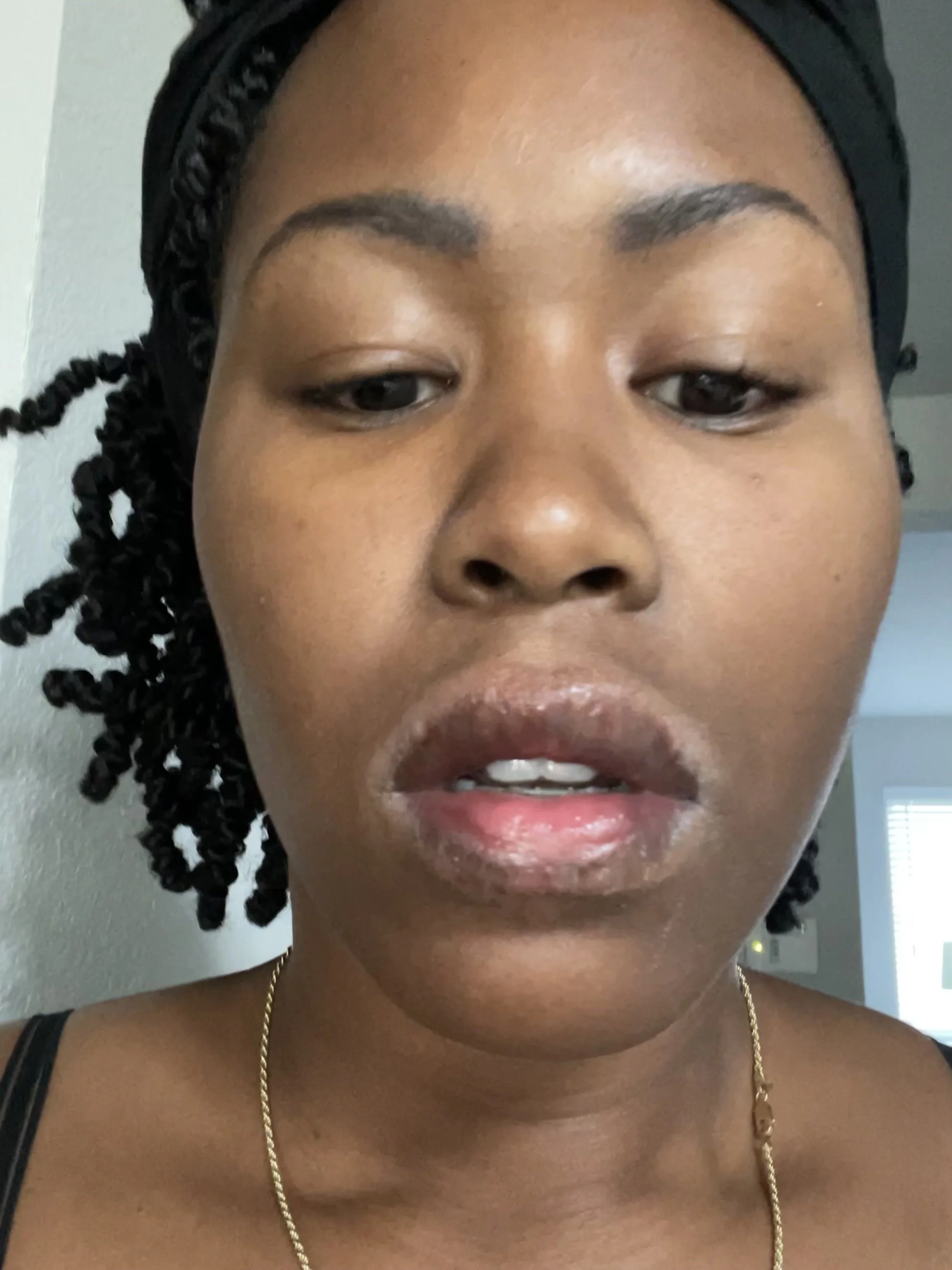 Close-up of a woman with twisted hair, wearing a black head covering and a gold necklace, standing indoors with a neutral expression.