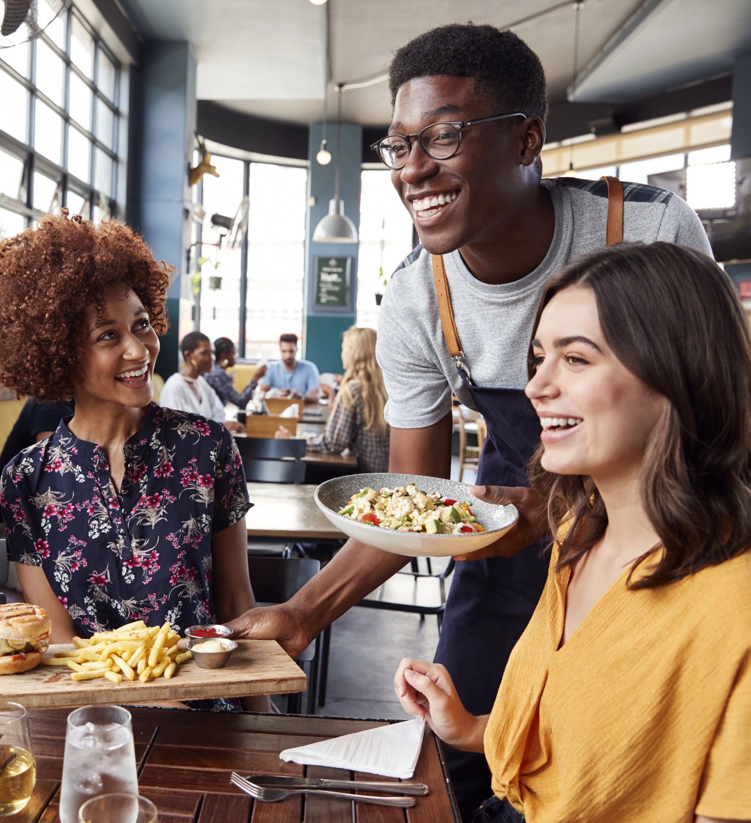 A smiling waiter serving salad to a woman seated at a restaurant table, with other diners in the background.