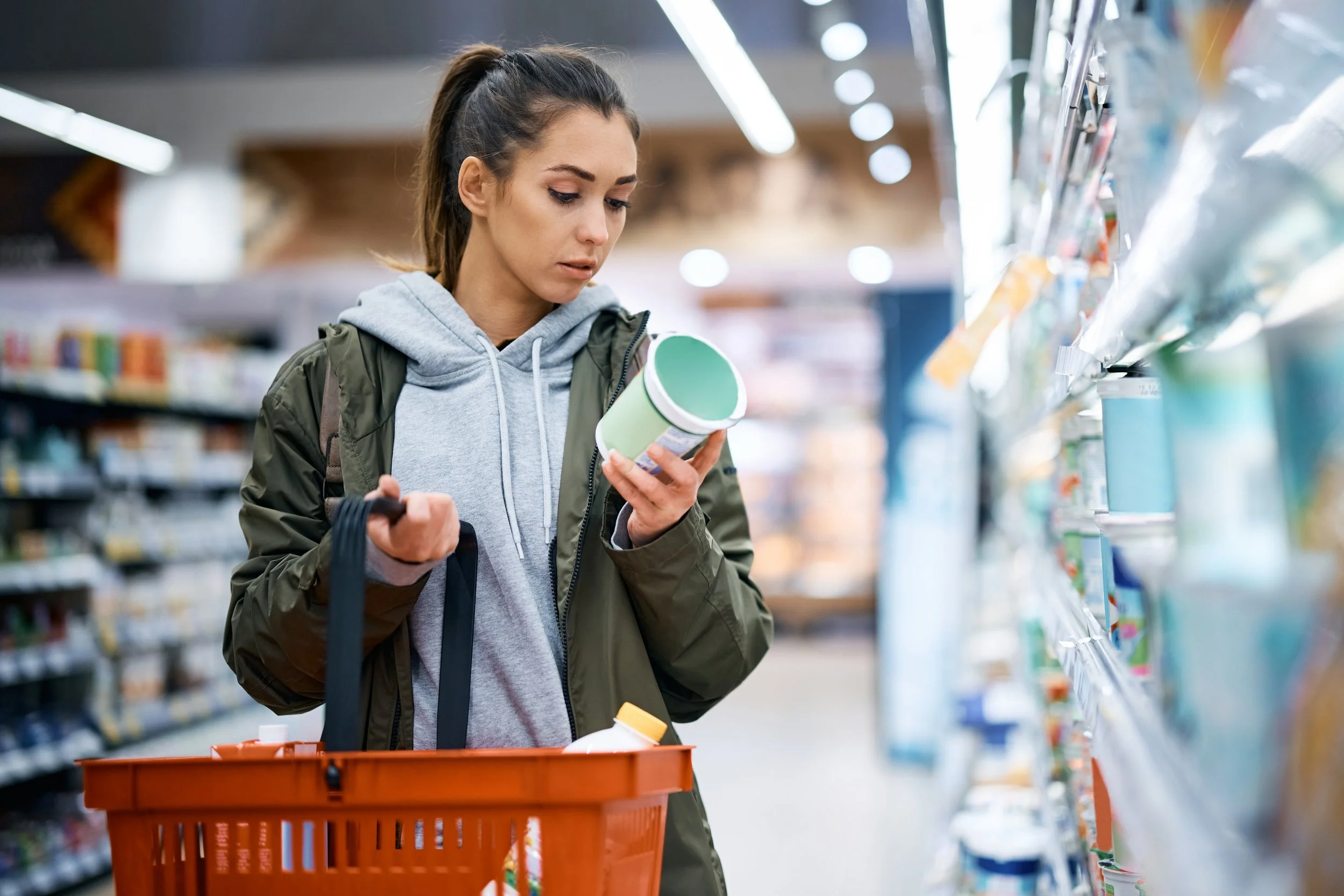 Young woman shopping in a grocery store aisle, holding a green container in her hand, with a shopping basket hanging from her arm.