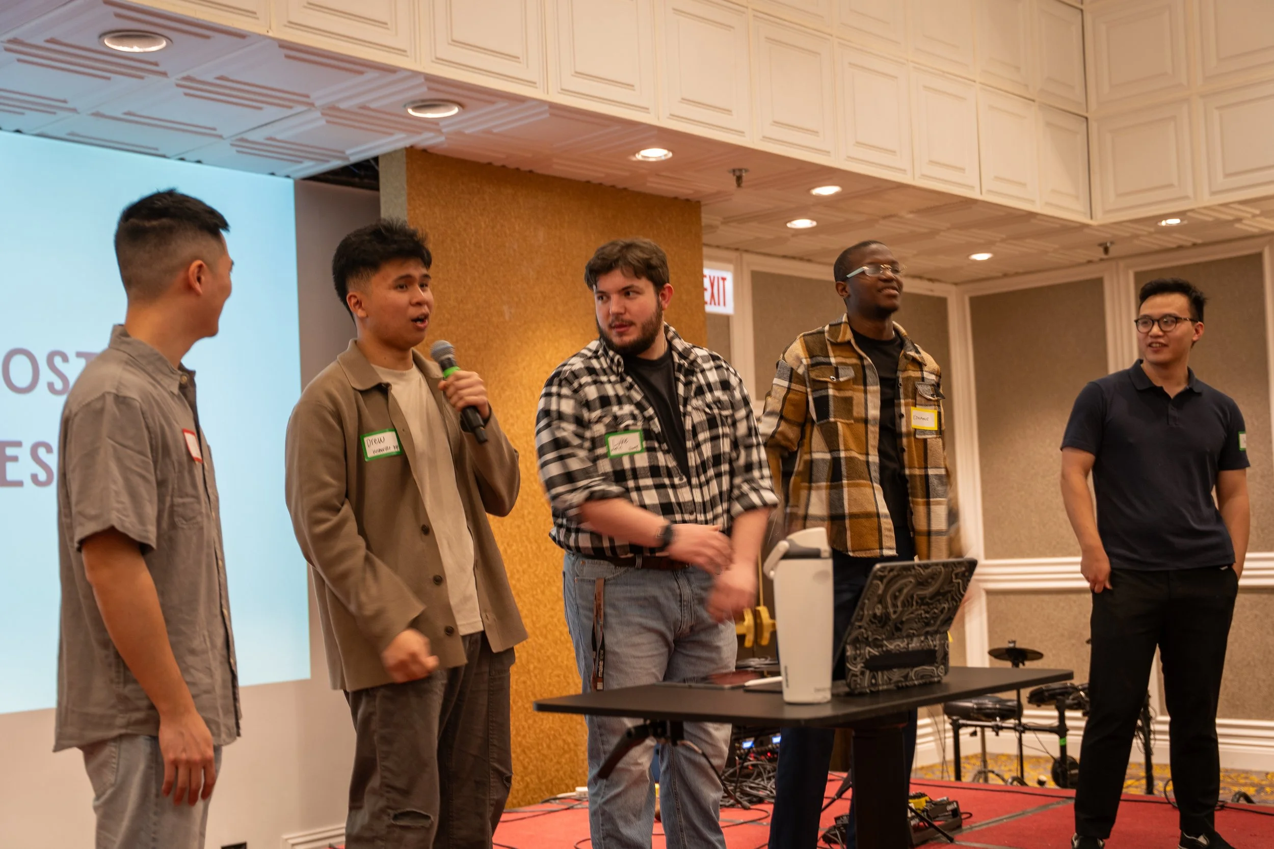 Five young men standing on a stage at a conference or event, with one speaking into a microphone and others listening. They are in front of a large screen, some wearing name tags, with electronic equipment on a table.