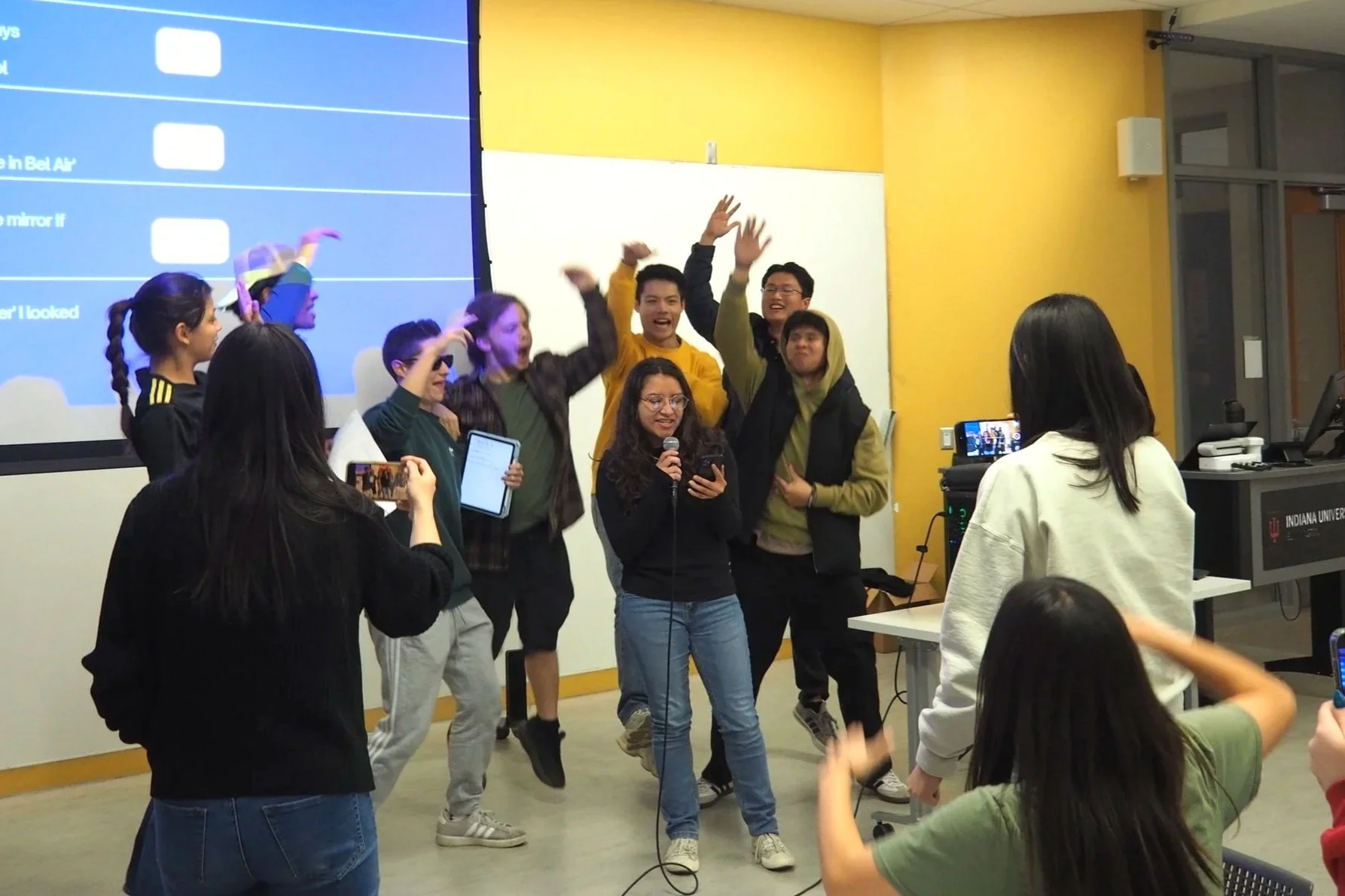 Group of students celebrating and singing in a classroom with a teacher or presenter holding a microphone, some students appear to be dancing or jumping, and others are recording the event on their phones.