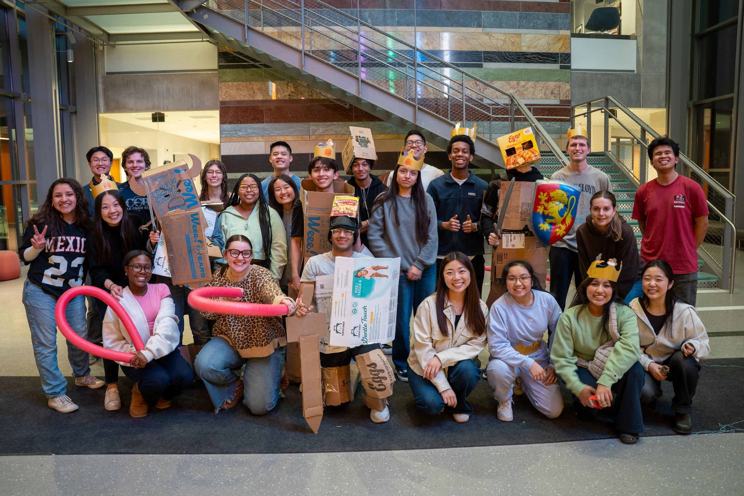Group of young people in an indoor setting, some wearing paper hats and holding cardboard boxes, smiling and posing for a group photo.