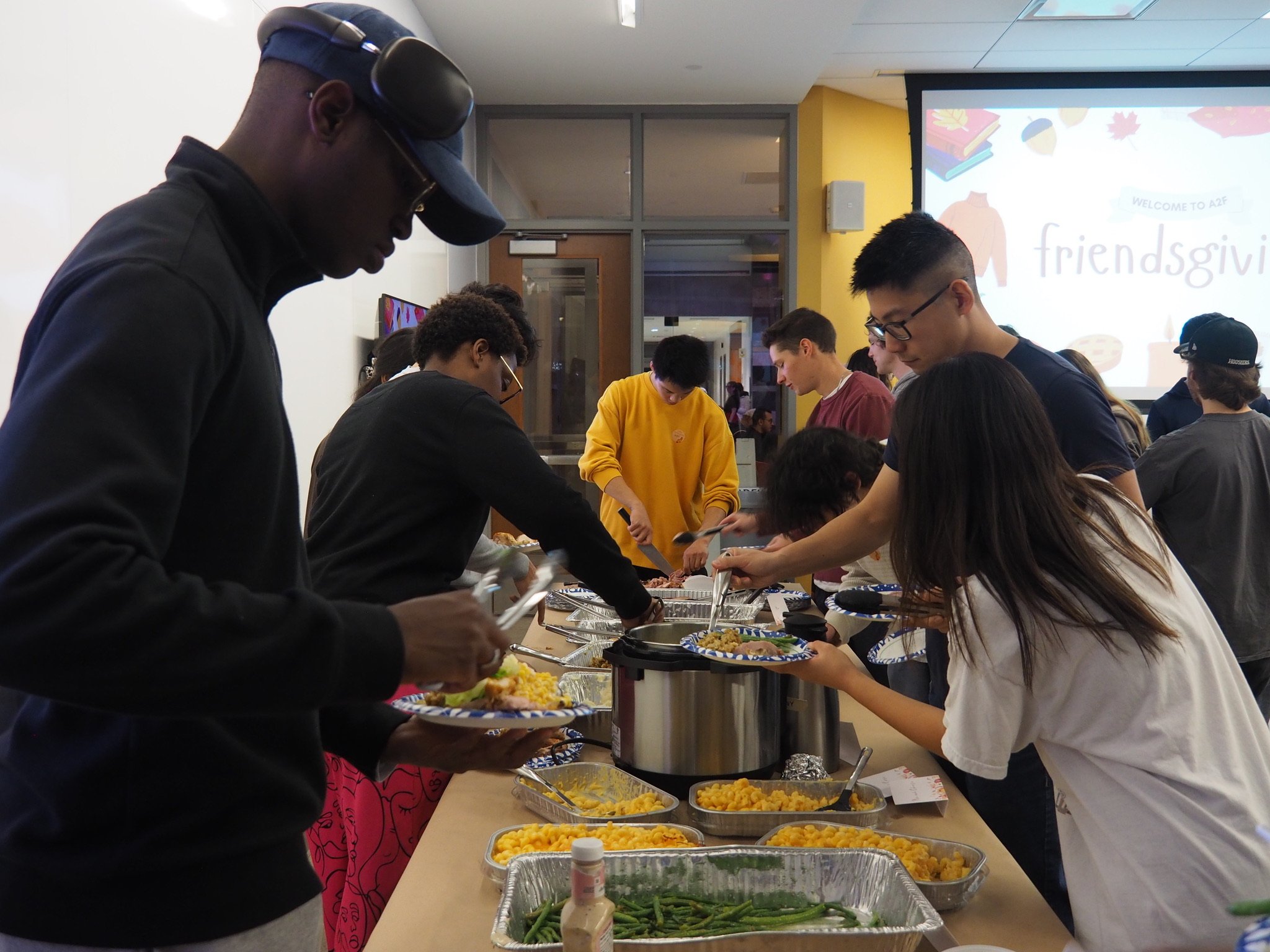 People serving themselves food at a buffet table during a gathering or event in a room with a large screen in the background that reads 'Friendsgiving'.