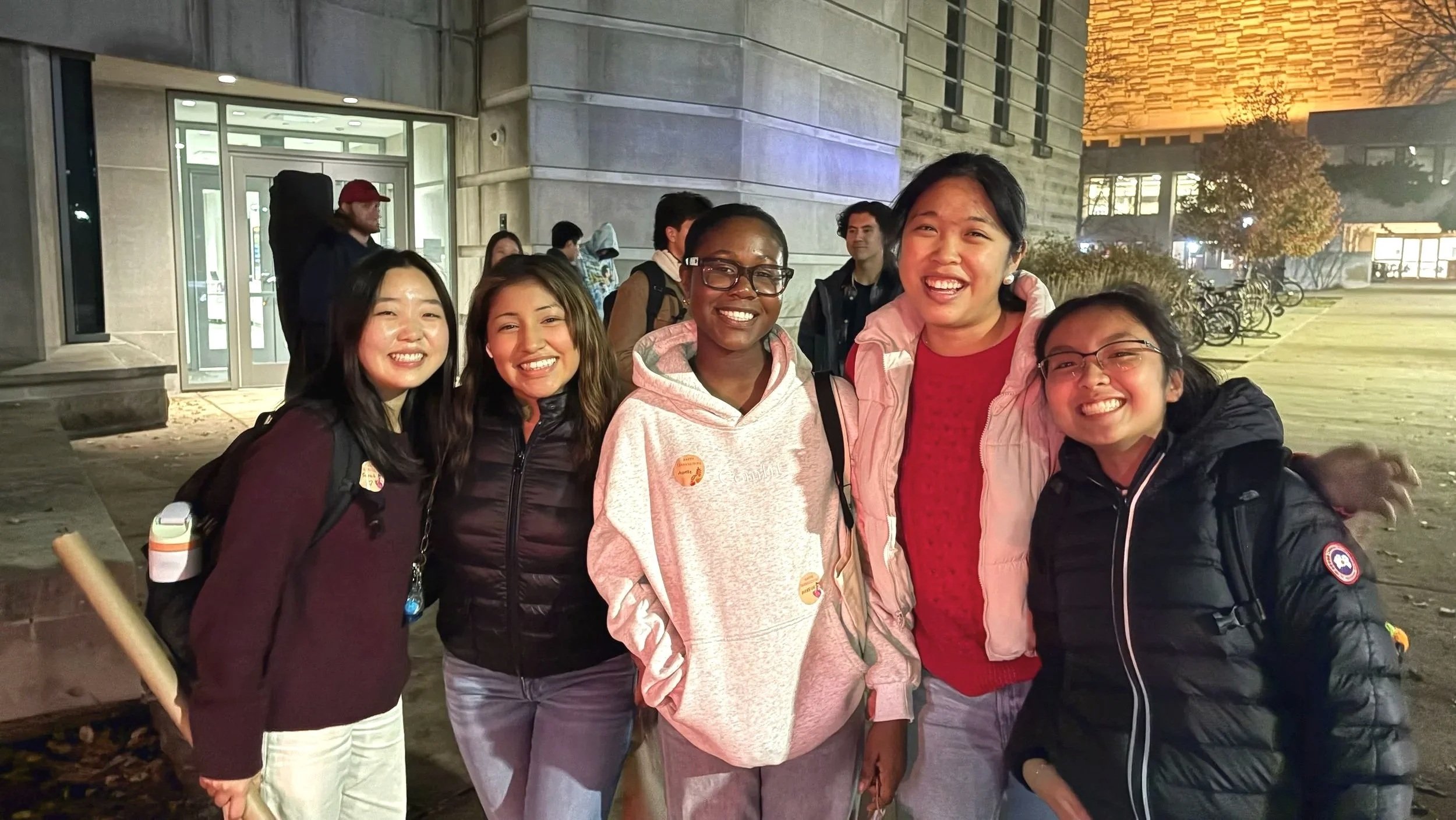 Five young women smiling and posing together outdoors at night, in front of a building with warm lighting and a few bicycles in the background.