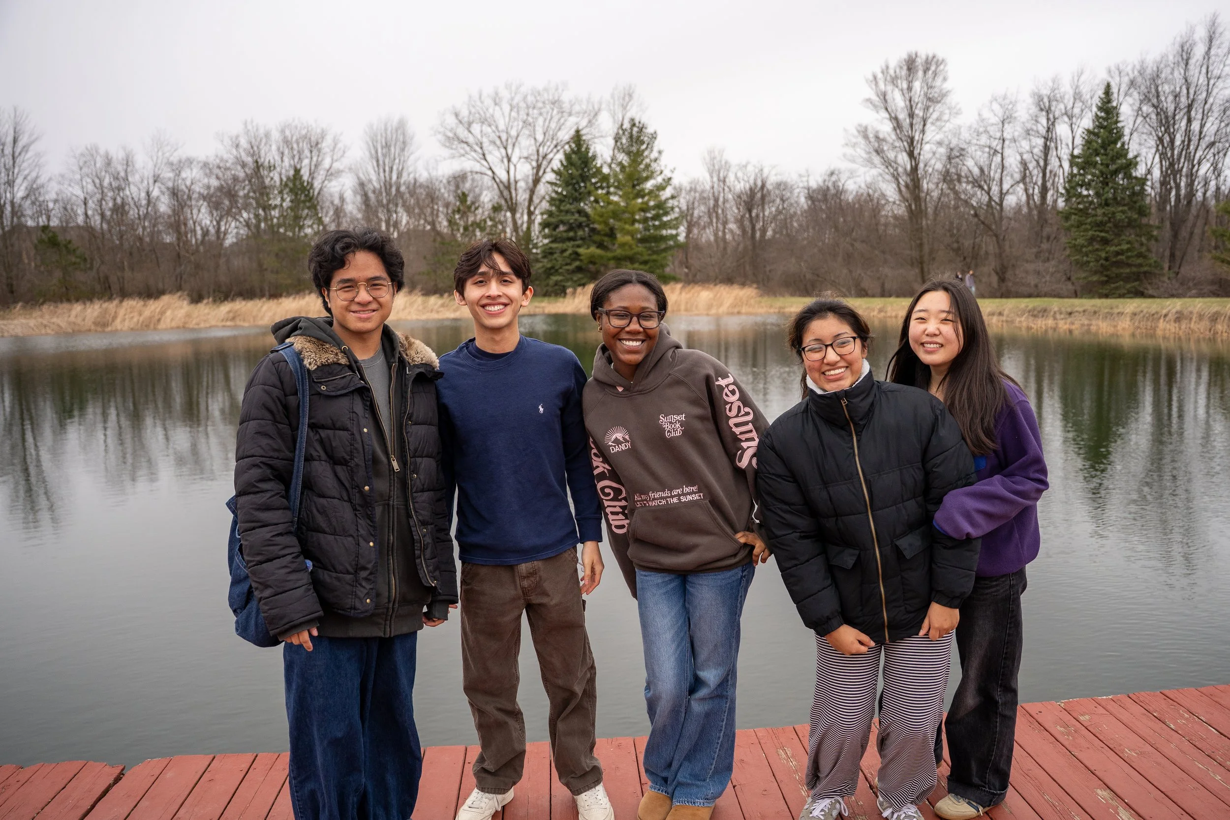 Group of five friends smiling on a wooden dock by a lake in a wooded area during overcast weather.