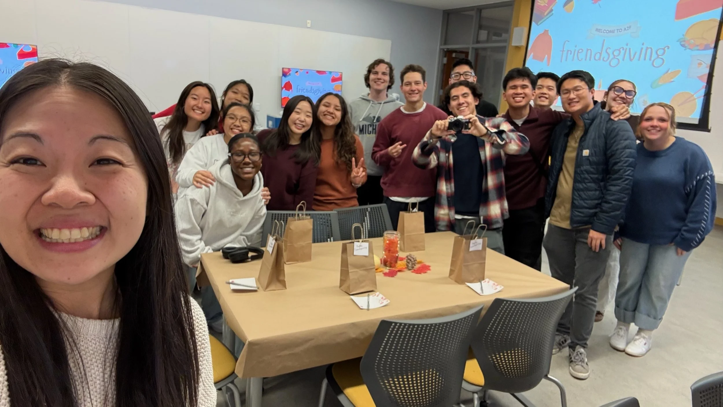 A group of diverse young adults gathered around a table for a friendsgiving celebration, with a woman taking a selfie in the foreground.
