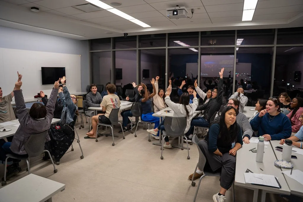 People sitting around tables in a classroom-like setting with many raising their hands, during a discussion or quiz, at night.