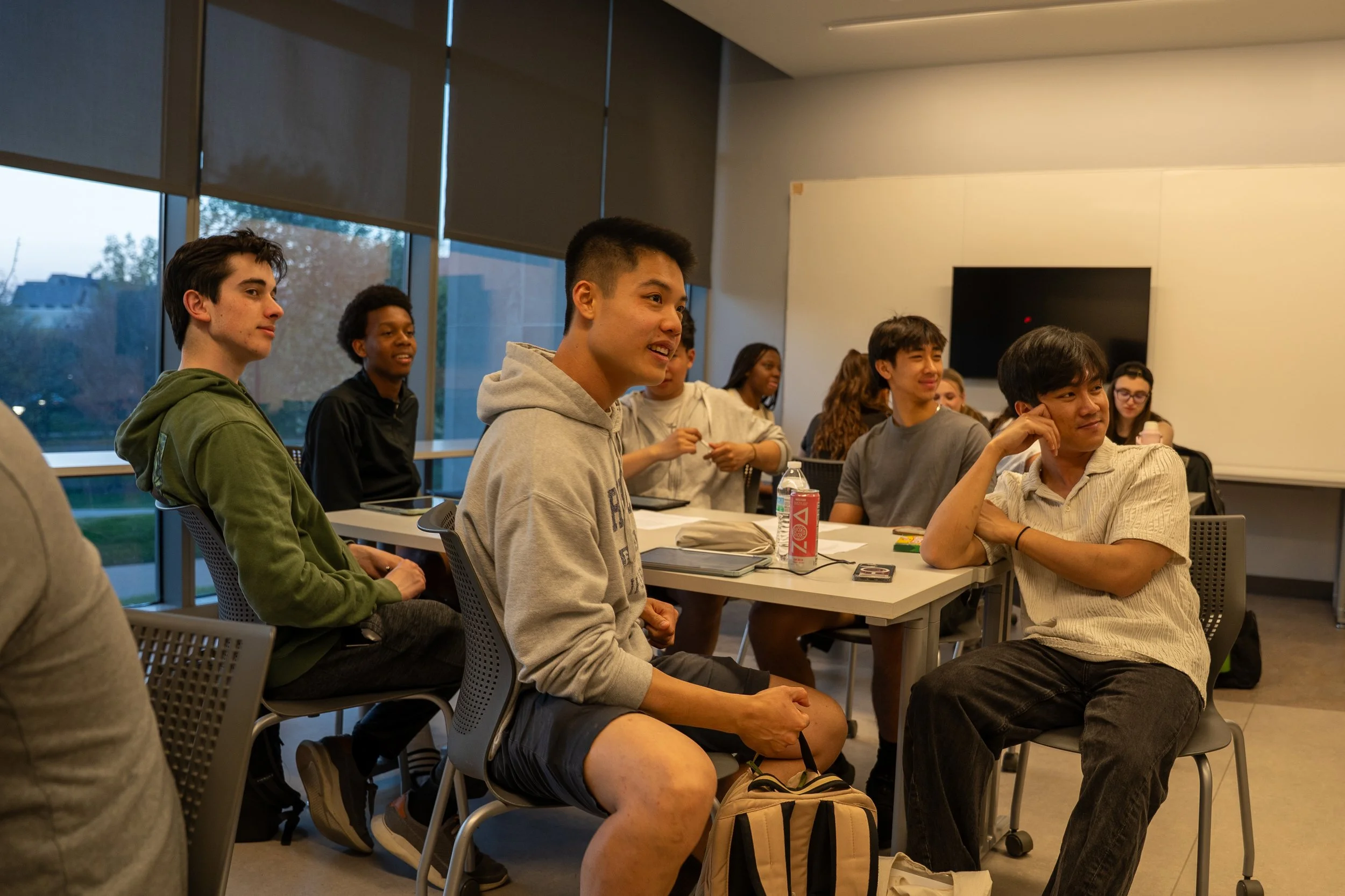 A group of diverse young adults sitting in a classroom or conference room, attentively listening and smiling during a presentation or discussion. The room has large windows with blinds and a modern interior.