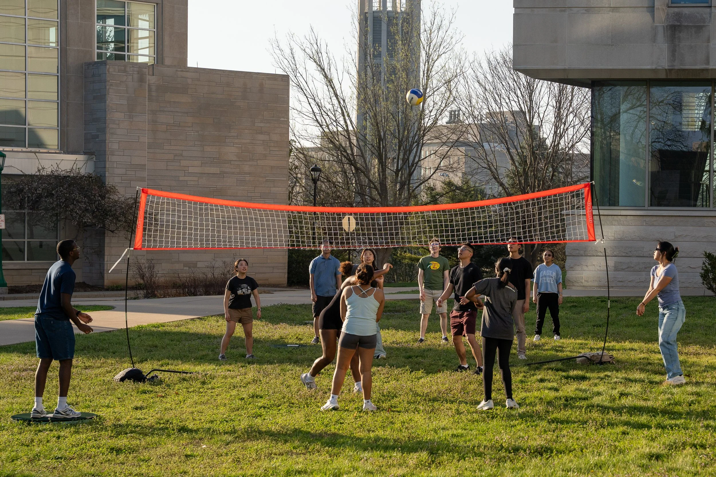 Group of people playing volleyball outdoors on a grassy area, with a net, in a city park or campus setting.