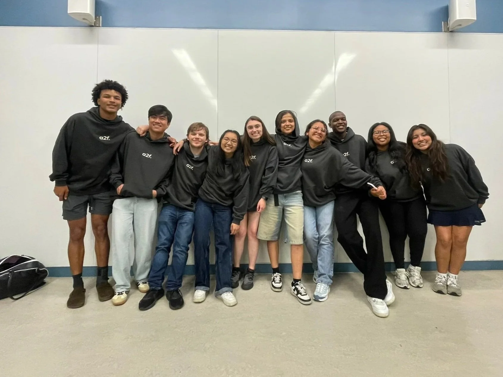 A diverse group of nine young people standing together in front of a plain wall, wearing black hoodies with a white logo, smiling and posing for a photo in an indoor setting.