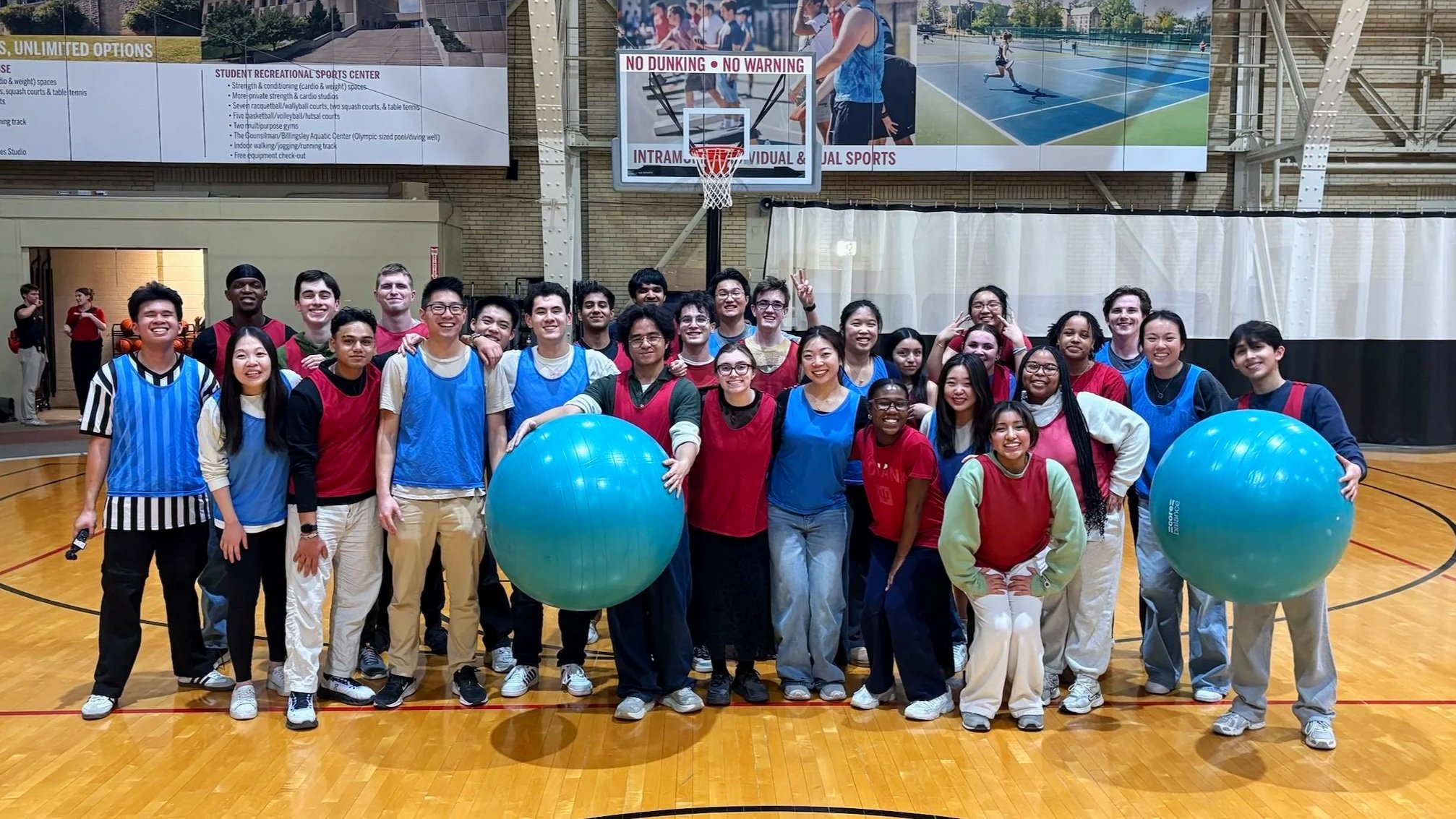 A group of diverse young people is gathered on a basketball court in a gymnasium, smiling at the camera. They are wearing colorful sports jerseys, with some holding large blue exercise balls. The background shows a basketball hoop, a large digital screen, and the high ceiling of the gym.