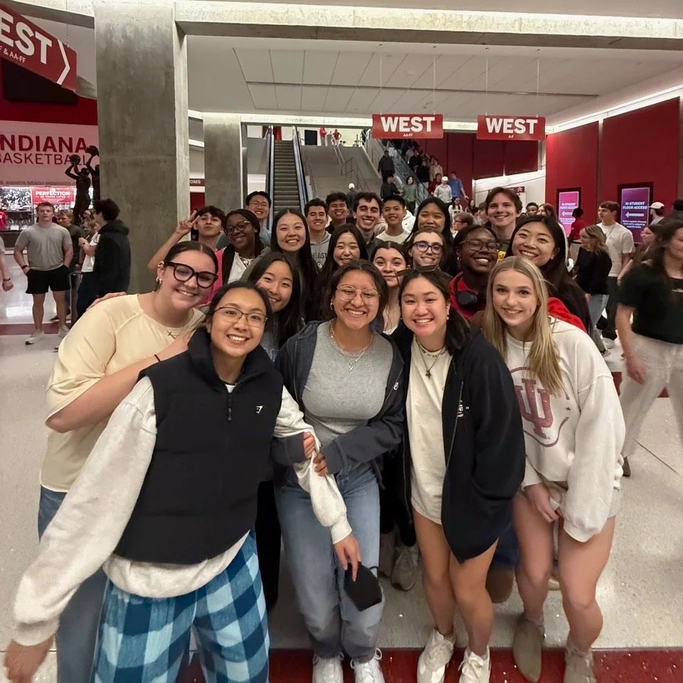 Group of college students smiling for a photo at an indoor stadium or arena entrance near escalators, with signs indicating the west entrance.
