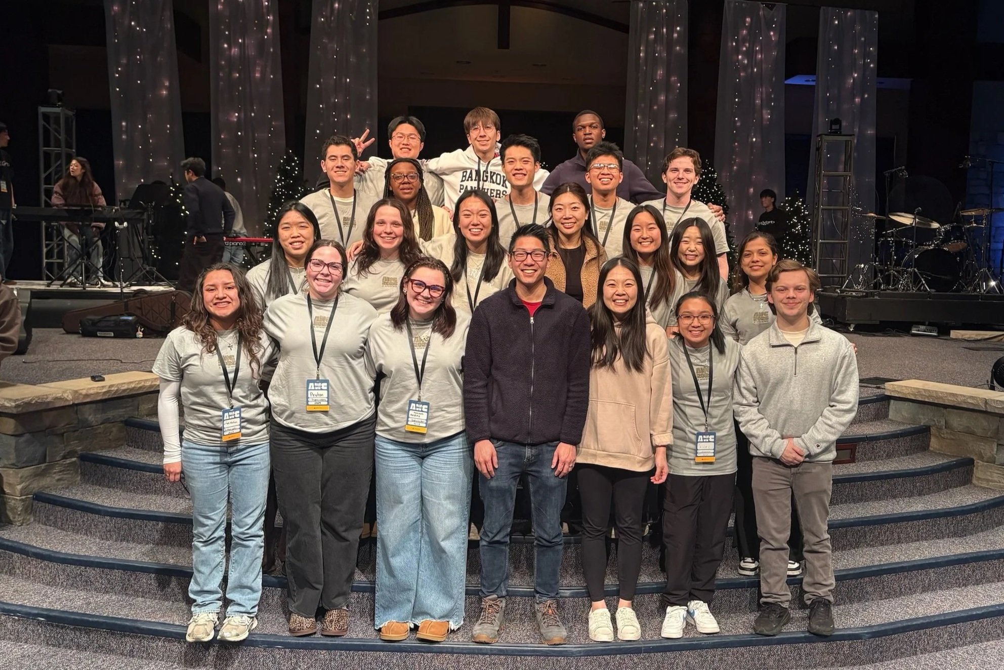 Group of young adults and a man posing on a stage with musical instruments and Christmas decorations in the background at an indoor event.