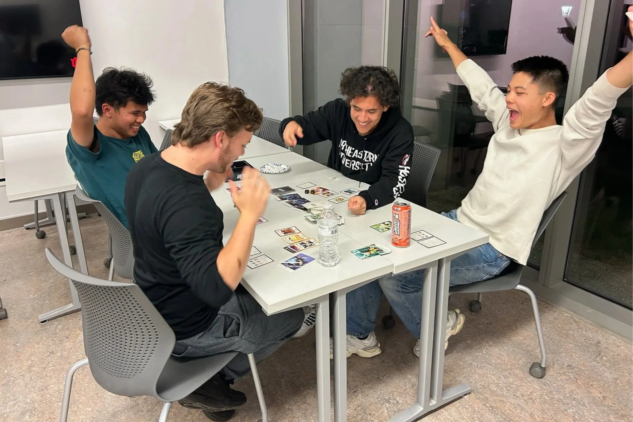 Four young men are sitting around a table, playing a card game and celebrating, with smiles and raised hands, in a modern room with glass doors and a TV in the background.