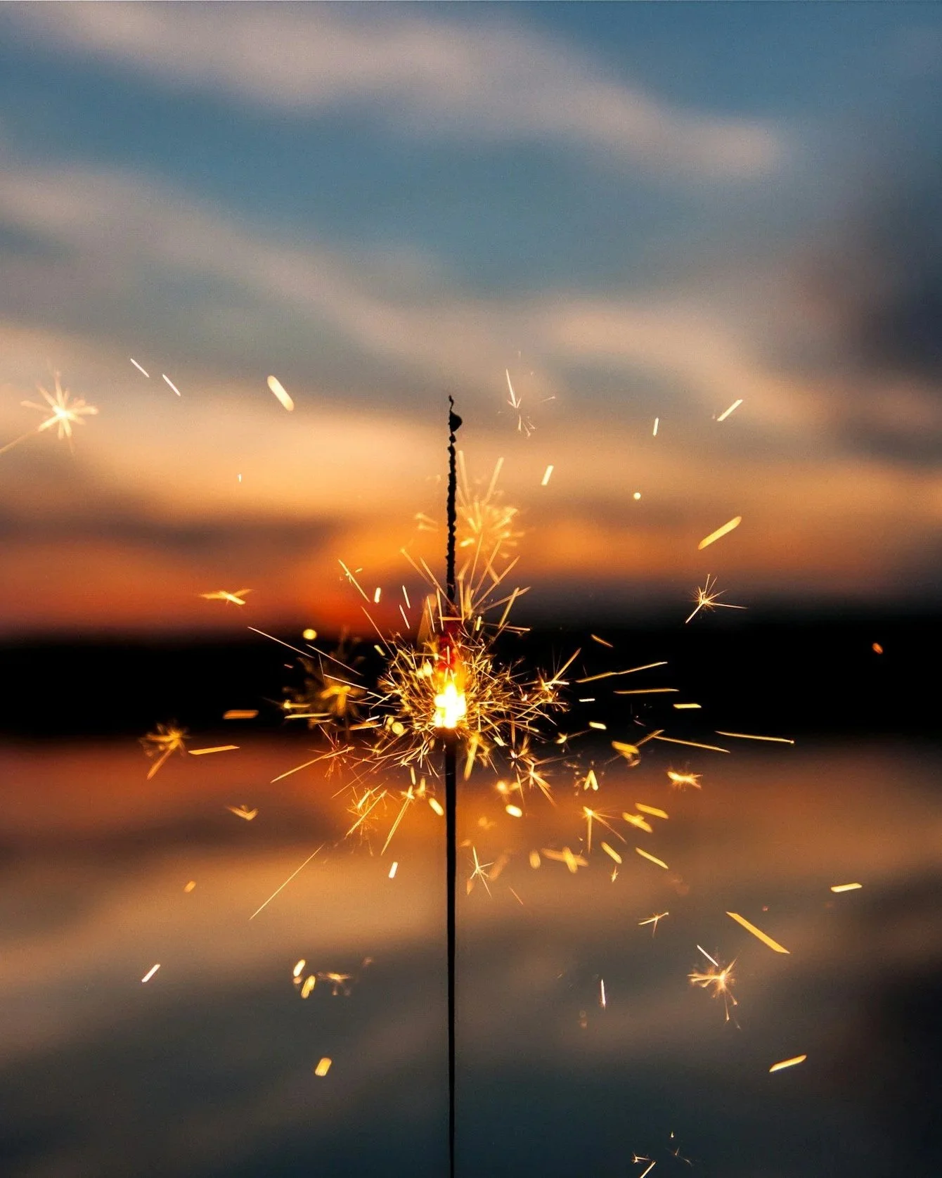 Sparklers burning on a stick at sunset over water with a colorful sky.