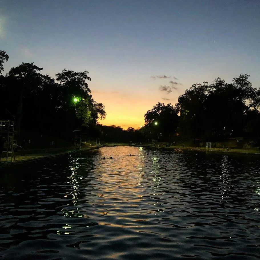 A pool at sunset with trees surrounding it and a few people swimming.