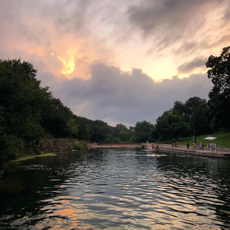 A peaceful river with a concrete path along the right side and people walking, some sitting under a white umbrella, during sunset with cloudy sky.