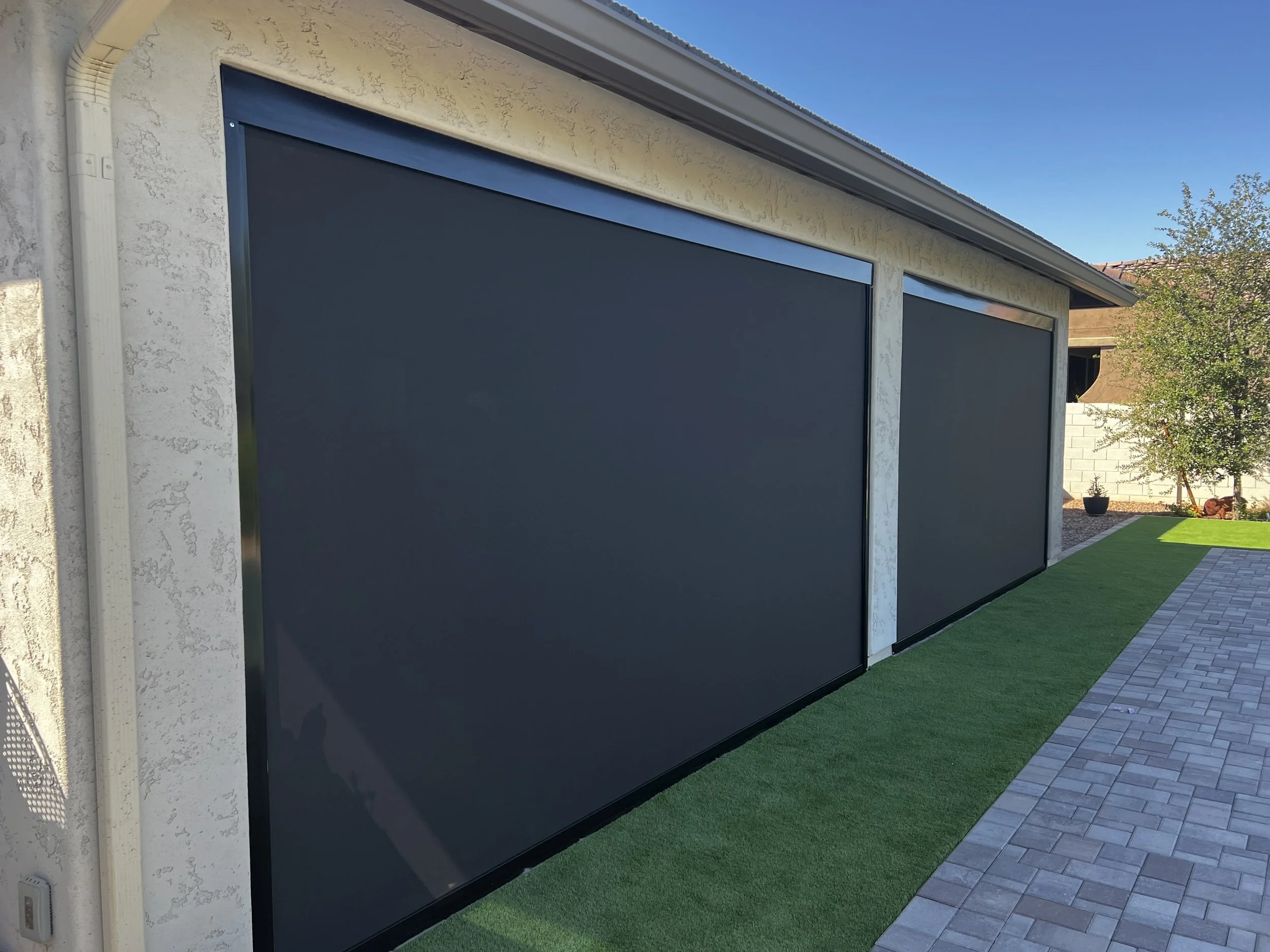 Two large black retractable window screens installed on the side of a beige stucco house with outdoor artificial grass and a brick patio leading to a yard with trees and plants.