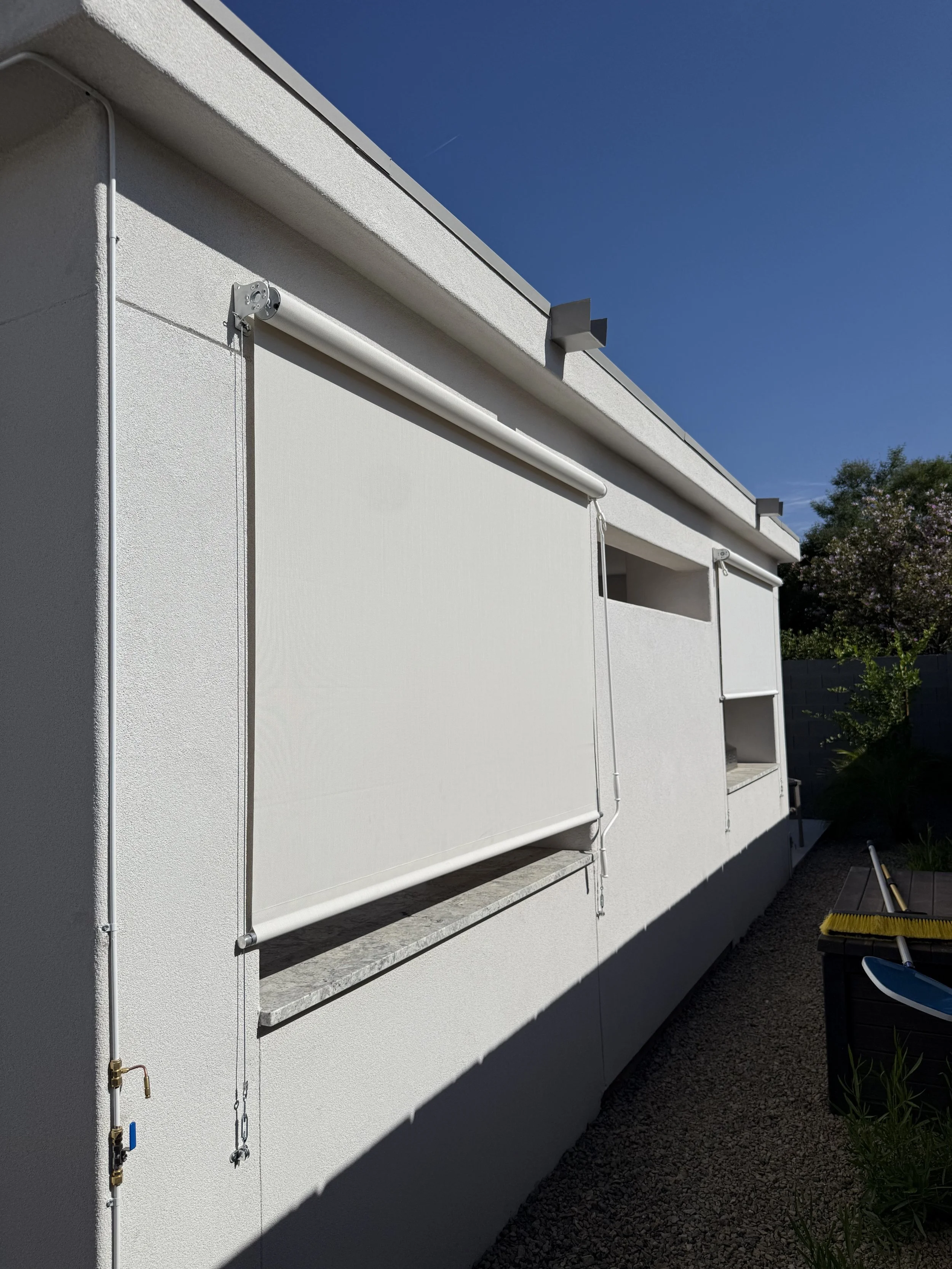 Exterior of a white building with closed white window blinds, a gravel ground, and a black storage container with gardening tools, under a clear blue sky.