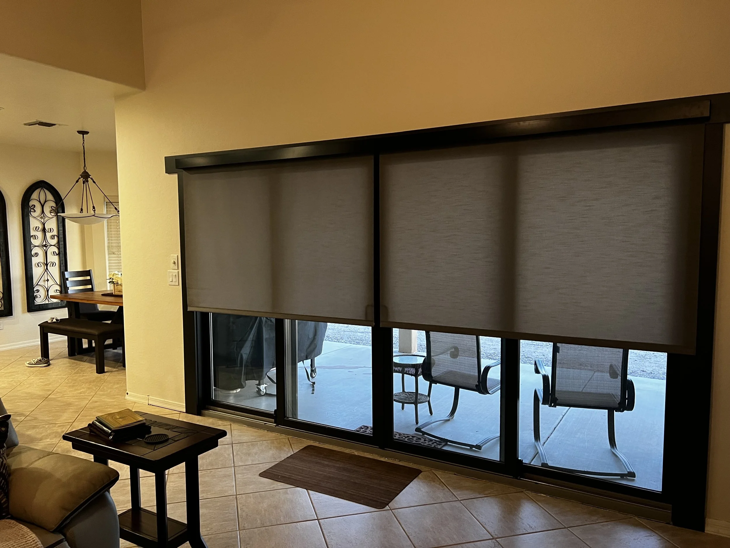 Living room with beige tiled floor, foldable window with beige shades covering the glass door, and a patio with chairs visible outside.