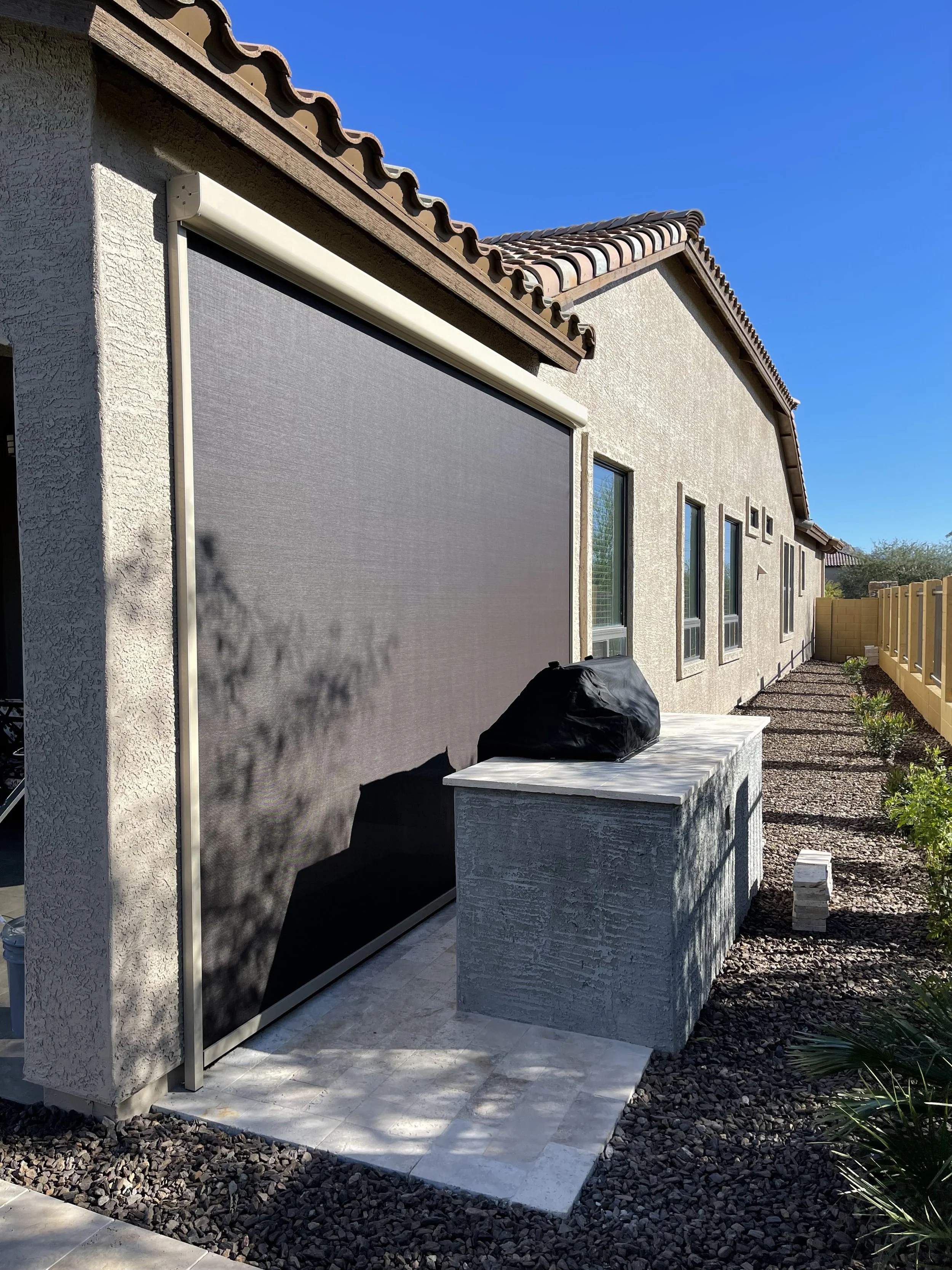 Outdoor patio area of a house with a large retractable screen, a barbecue covered with a black cover, and a small ledge or counter, illuminated by bright sunlight under a clear blue sky.
