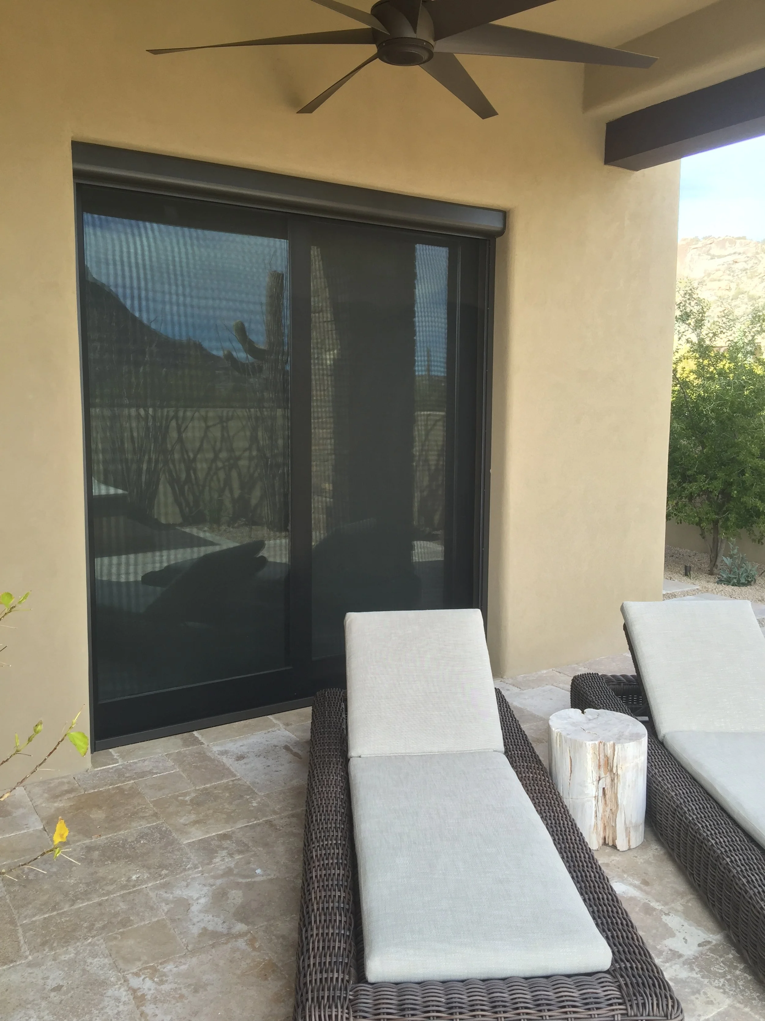 Outdoor patio with two chaise lounges, a wooden side table, sliding glass door, ceiling fan, and desert landscape in the background.
