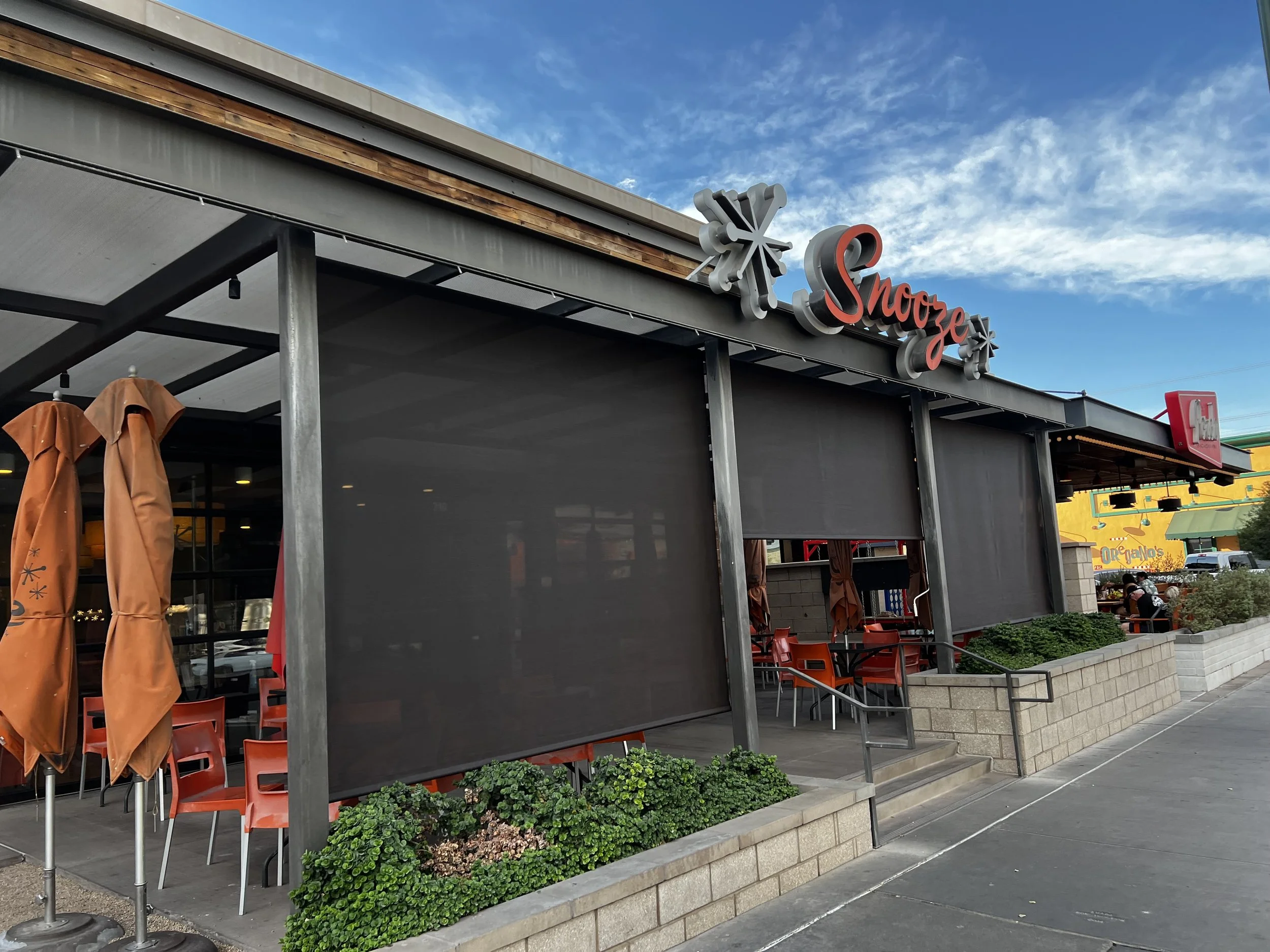 Outside of a restaurant named Snooze with orange chairs, umbrellas, planters, and a blue sky.