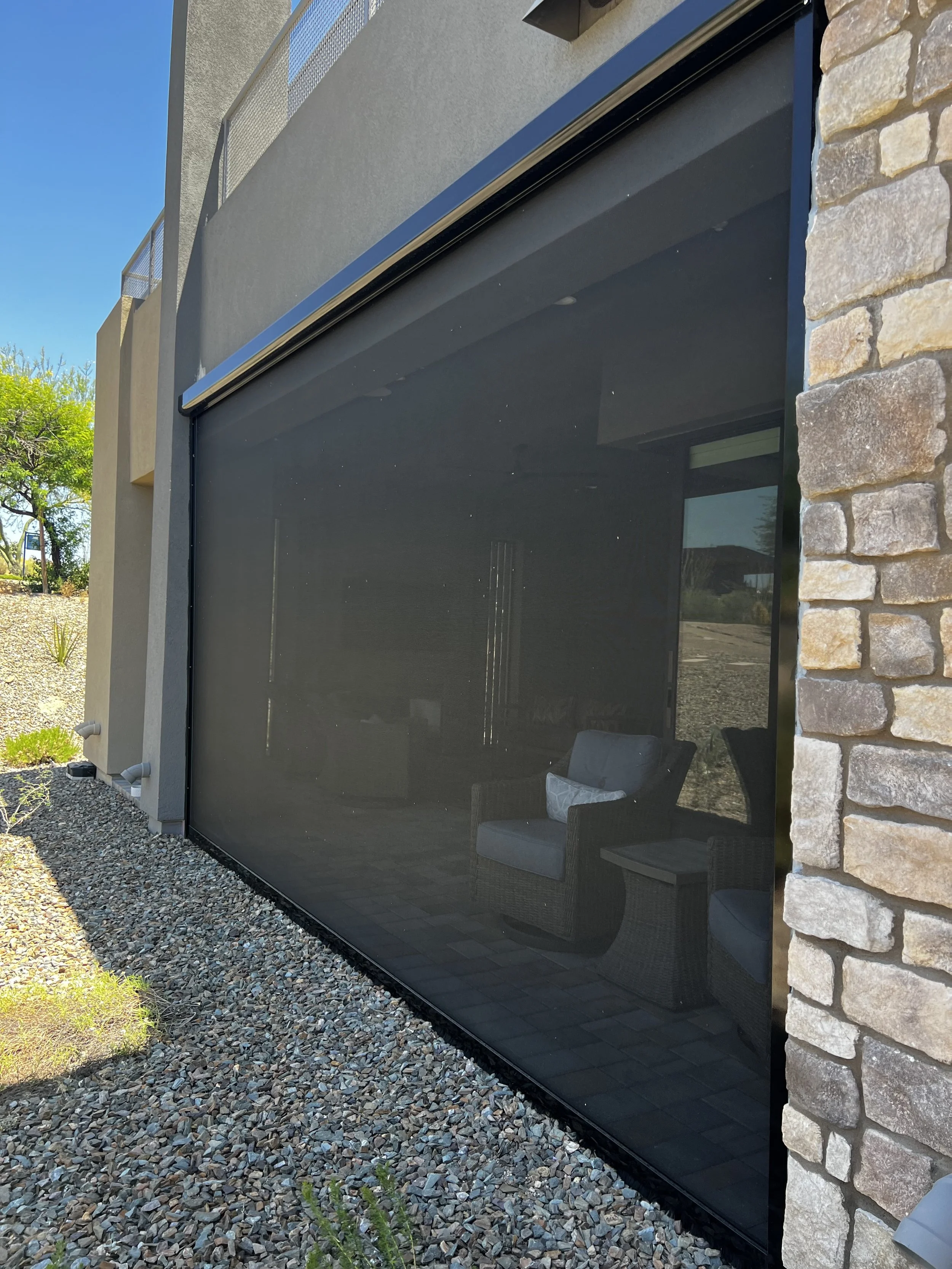 View of a house with a large black screen enclosure, patio furniture inside, gravel ground, stone wall on the right, and a tree with green leaves under a clear blue sky.