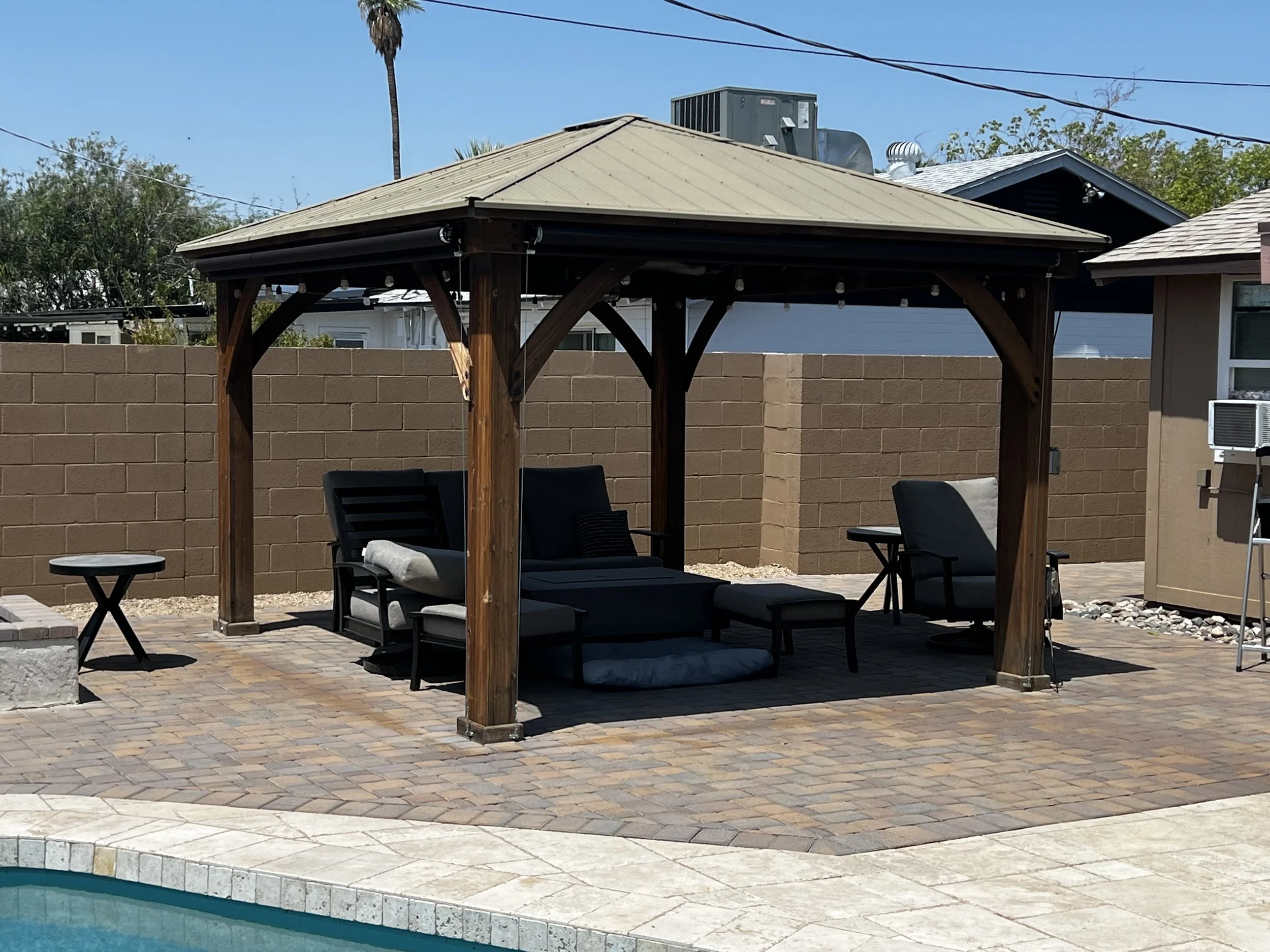 Backyard swimming pool area with a wooden gazebo, outdoor seating including lounge chairs, and a brick paver patio, with a brown cinder block wall in the background.