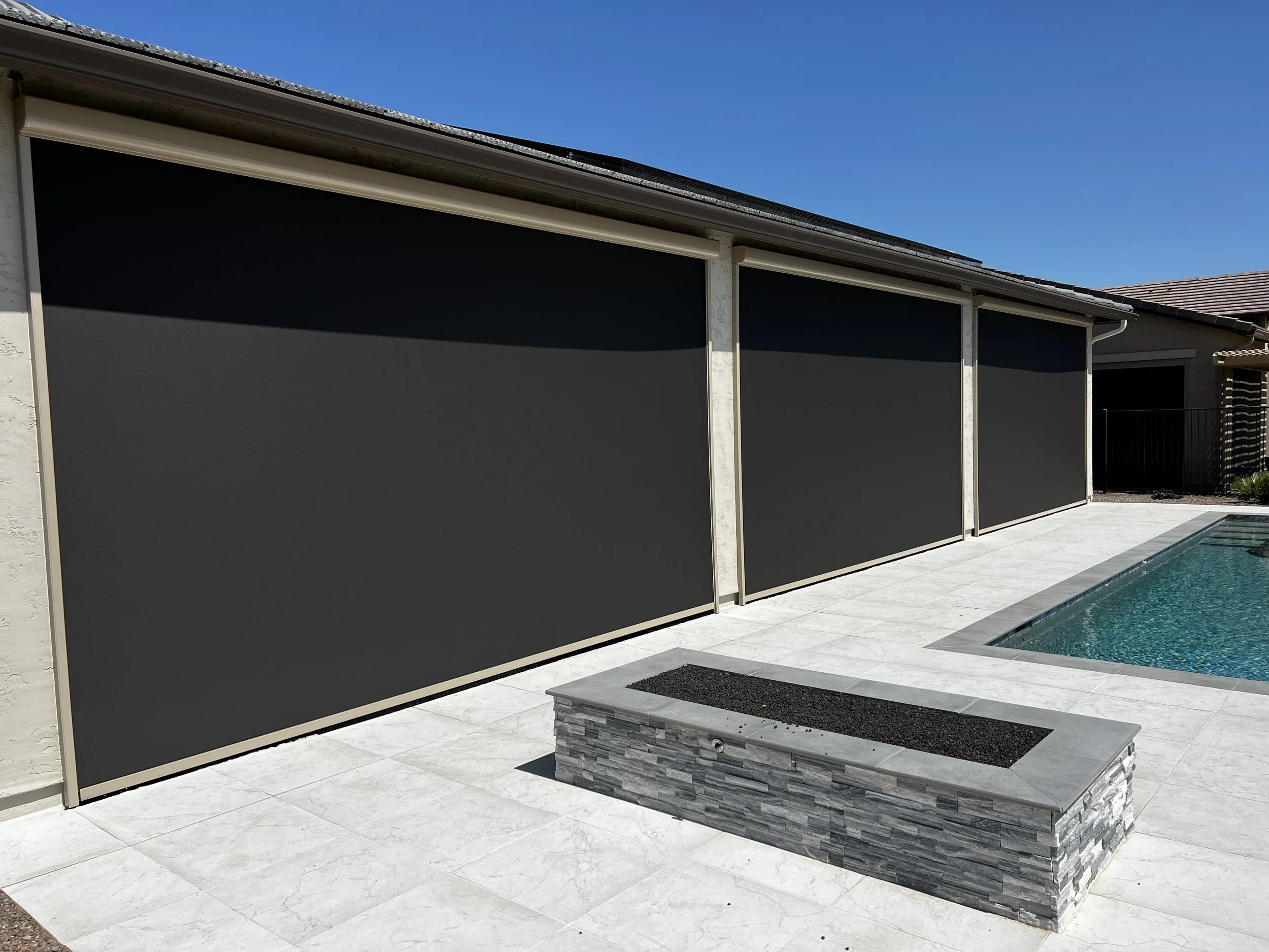 Backyard with a swimming pool, stone planter box, and screen shades on a patio, under a bright blue sky.