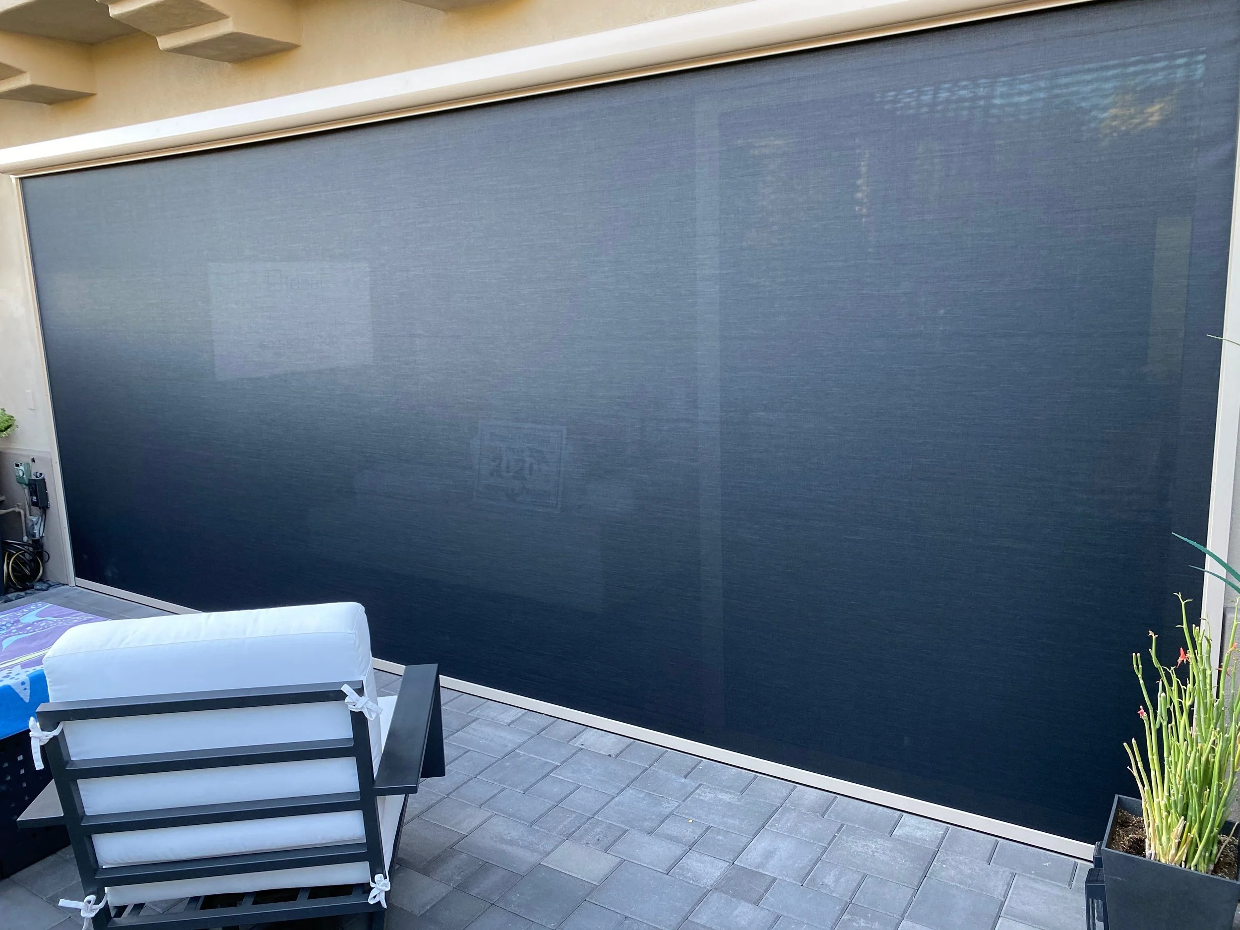 A large blue outdoor retractable screen or shade on a patio with a metal chair, a small table with a blue cover, and potted plants on a tiled floor.