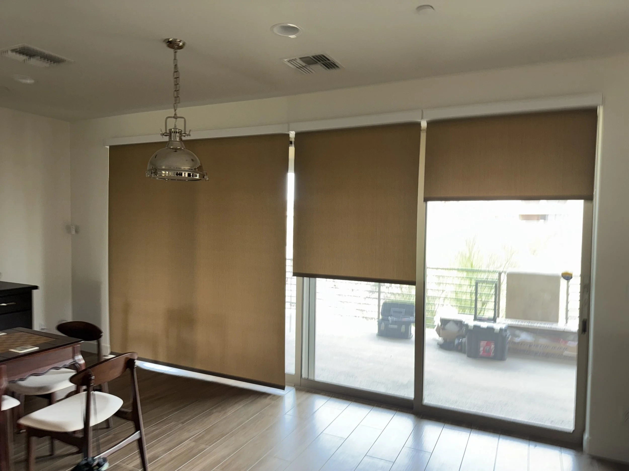 Living room with brown window blinds covering large sliding glass doors, a wooden dining table with white chairs, and a hanging metallic pendant light.