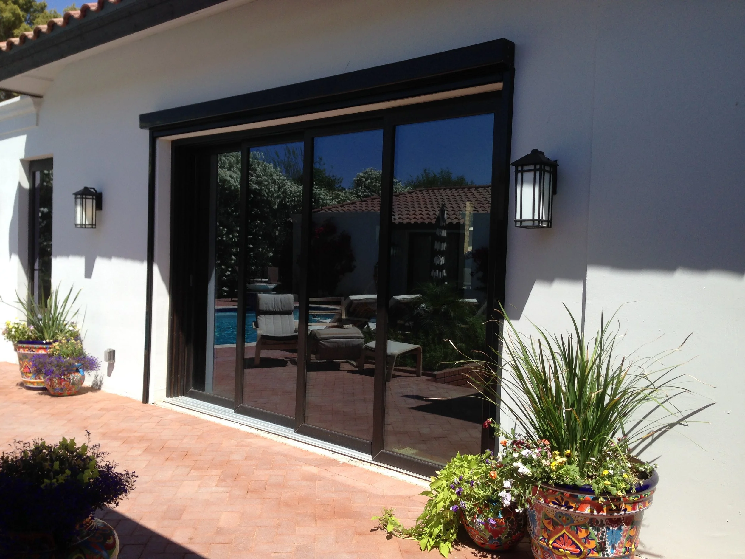 View of a patio with potted plants, a sliding glass door, and outdoor furniture reflecting in the glass, with a pool visible beyond.