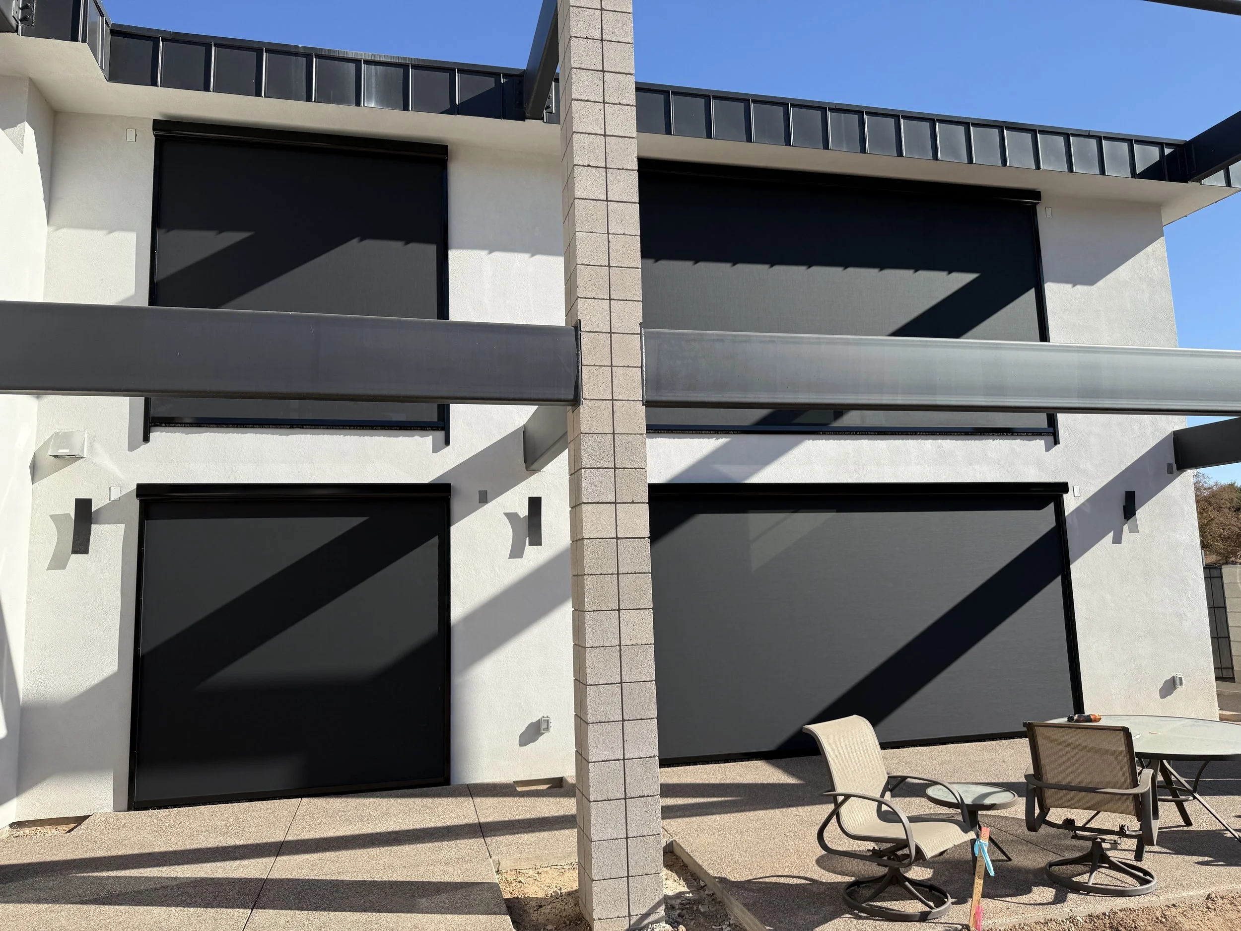 Modern two-story building with black roller shades on windows, outdoor patio furniture, and a clear blue sky.