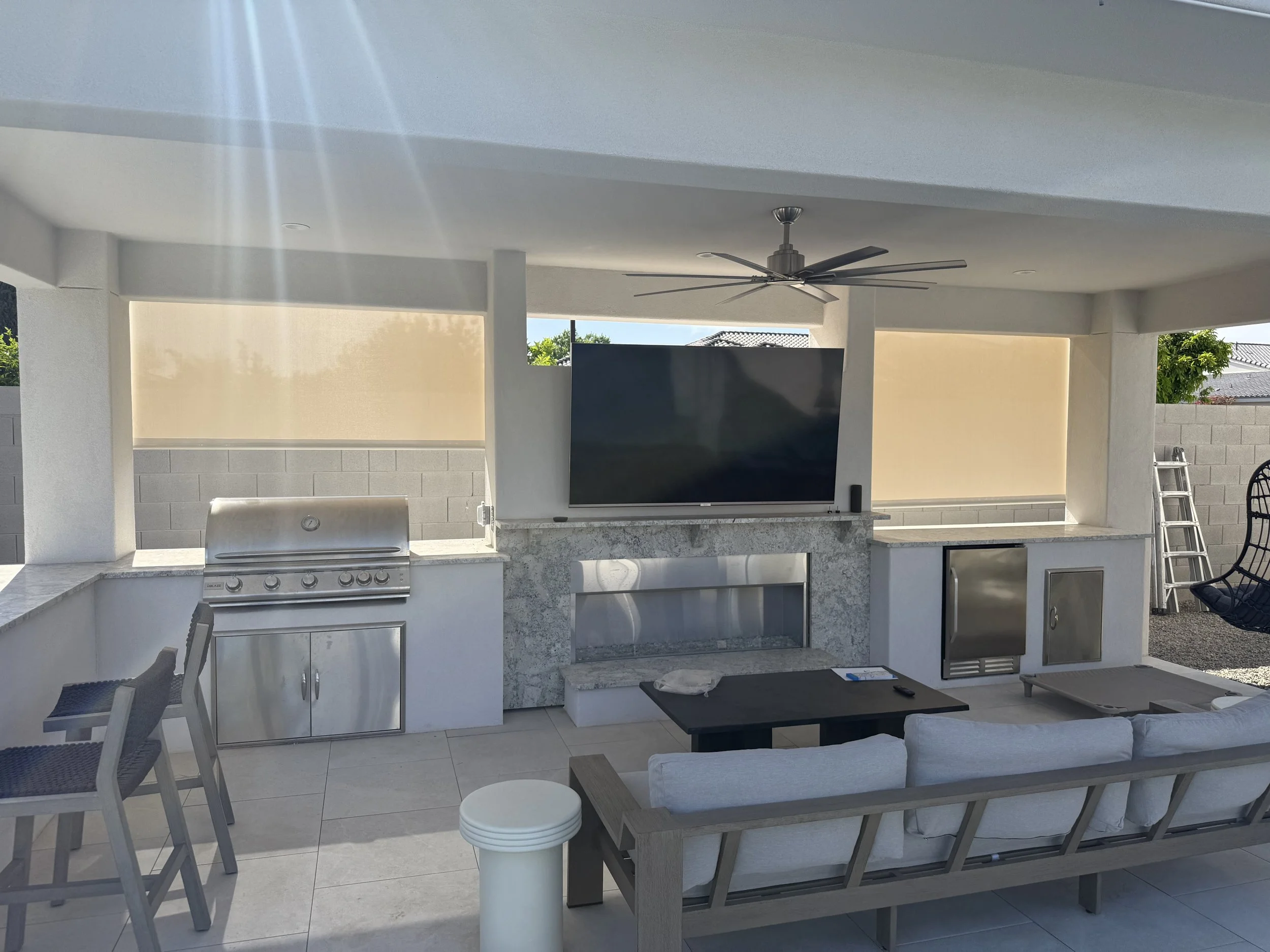 Outdoor patio with a built-in grill, a large flat-screen TV, a ceiling fan, a couch facing the TV, a small table, and chairs, with a ladder in the corner and a dog bed on the ground.