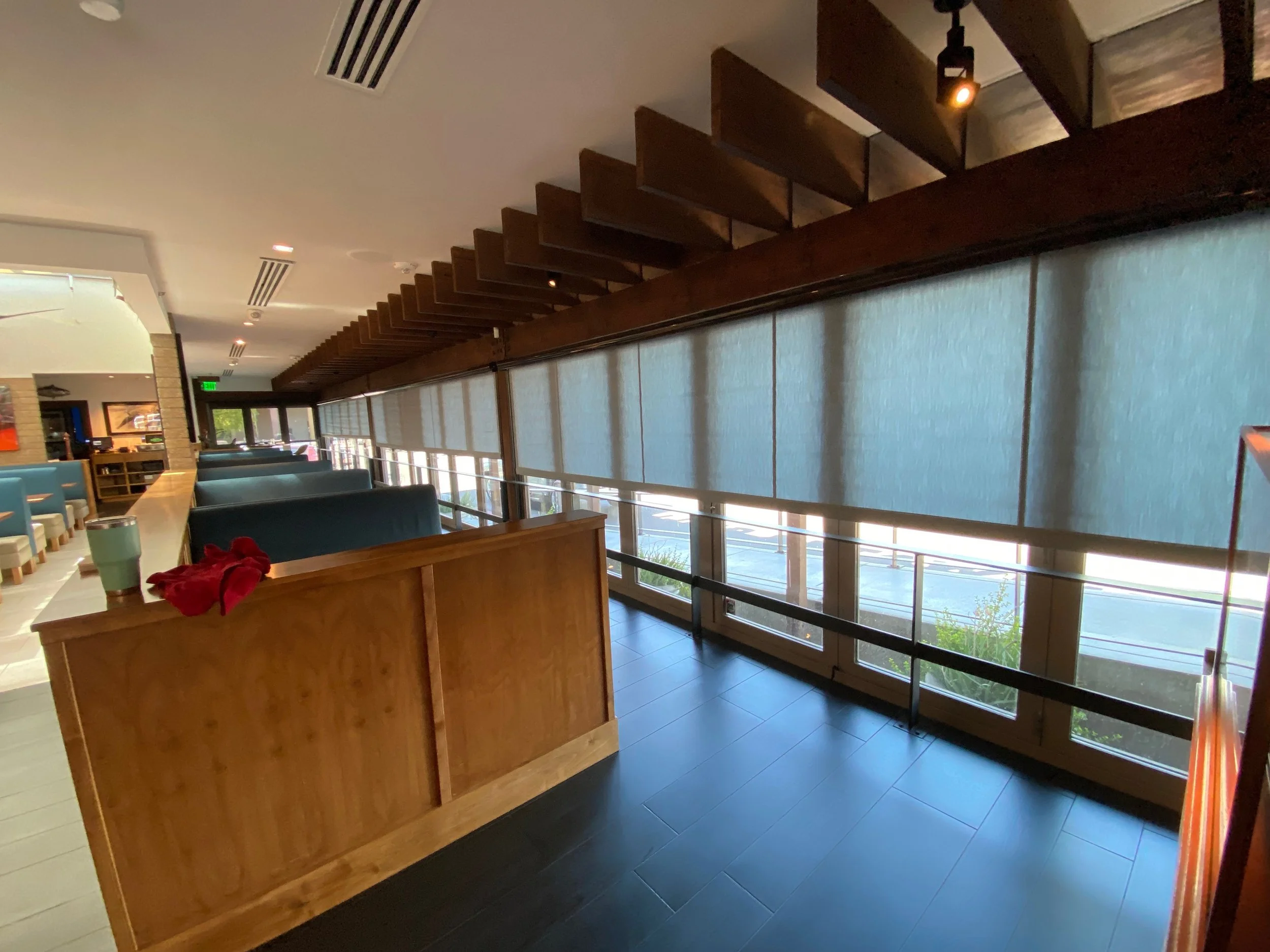Interior of a modern restaurant with booths, large windows with roller shades, wooden accents, and subtle lighting.