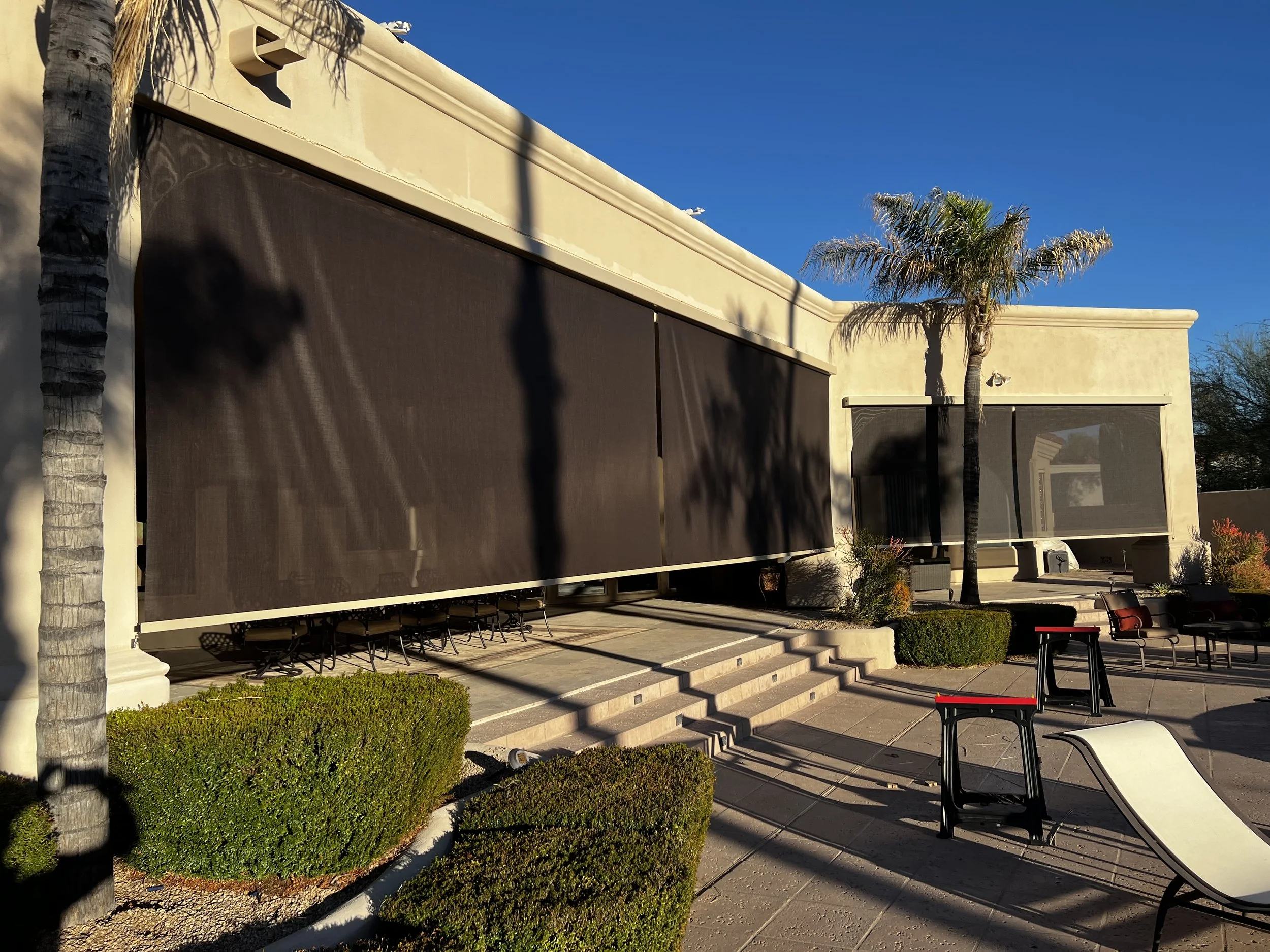 Modern building with beige walls, large black sun shades, and outdoor seating area with palm trees and bushes.