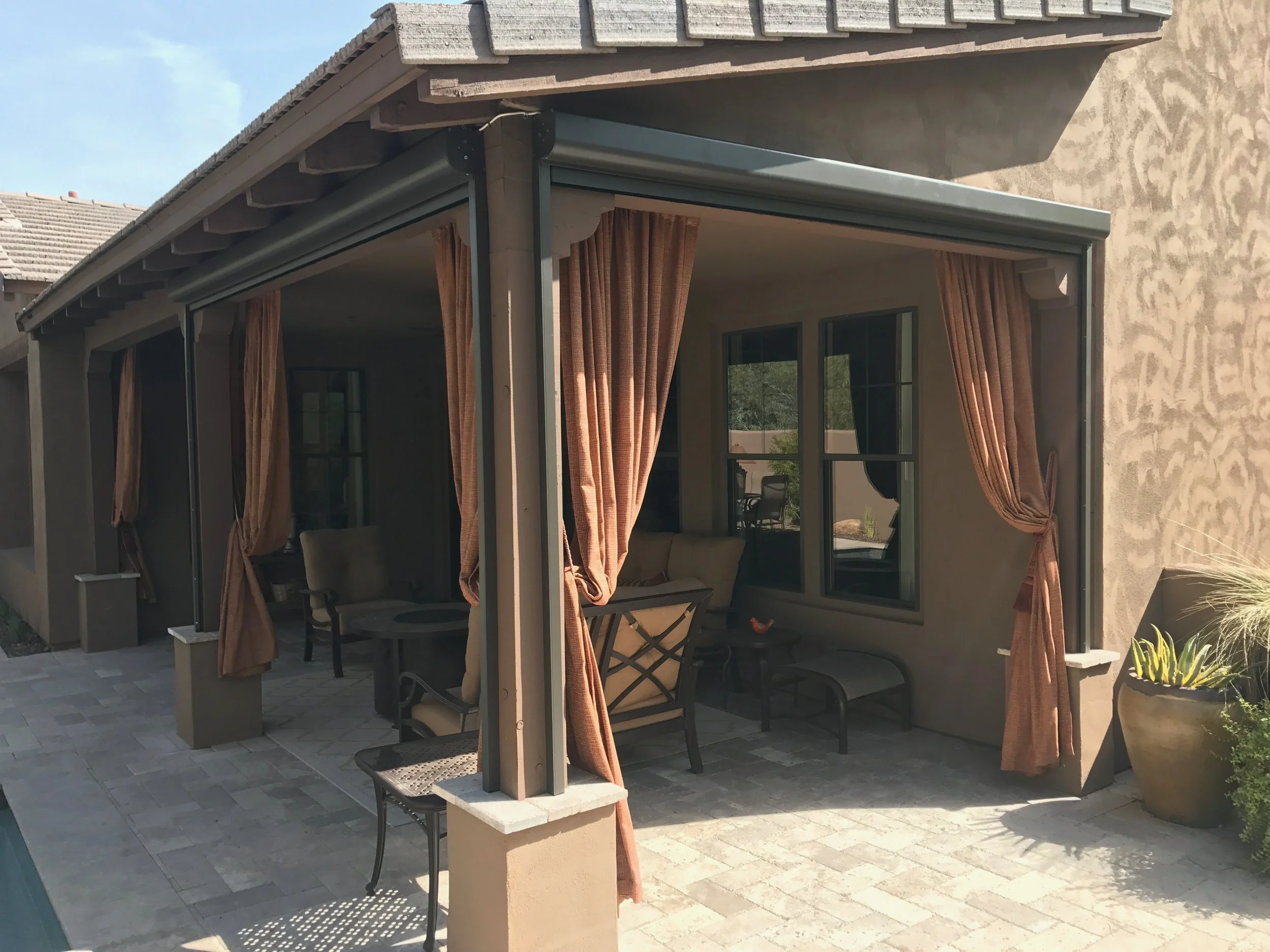 Outdoor patio with brown curtains, seating area with chairs and a small table, and windows on a beige stucco house.