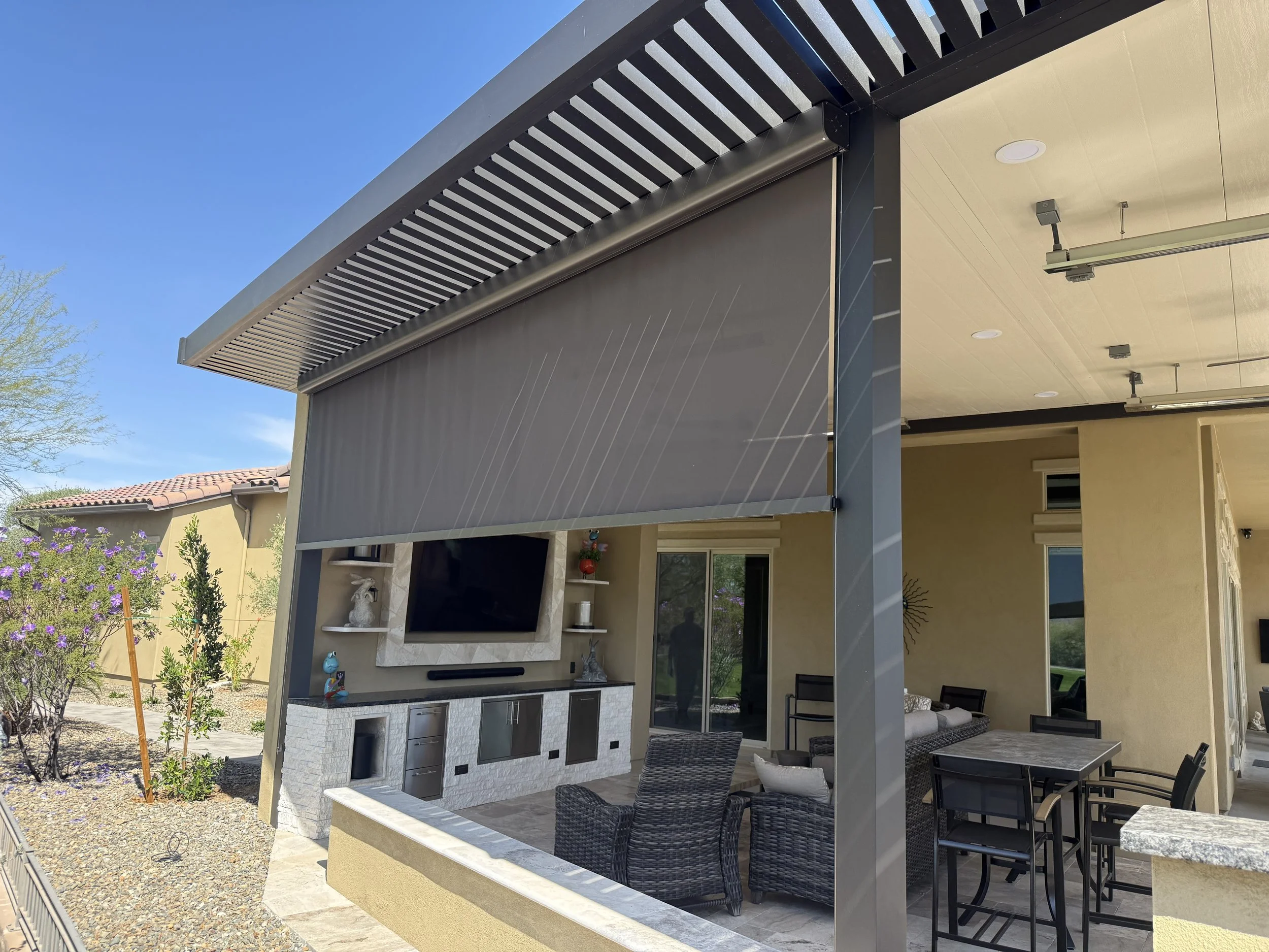 Outdoor patio with a ceiling mounted TV, wicker seating, a dining table with chairs, and a retractable shade, attached to a beige house with sliding glass doors, under a pergola with black beams and a blue sky.