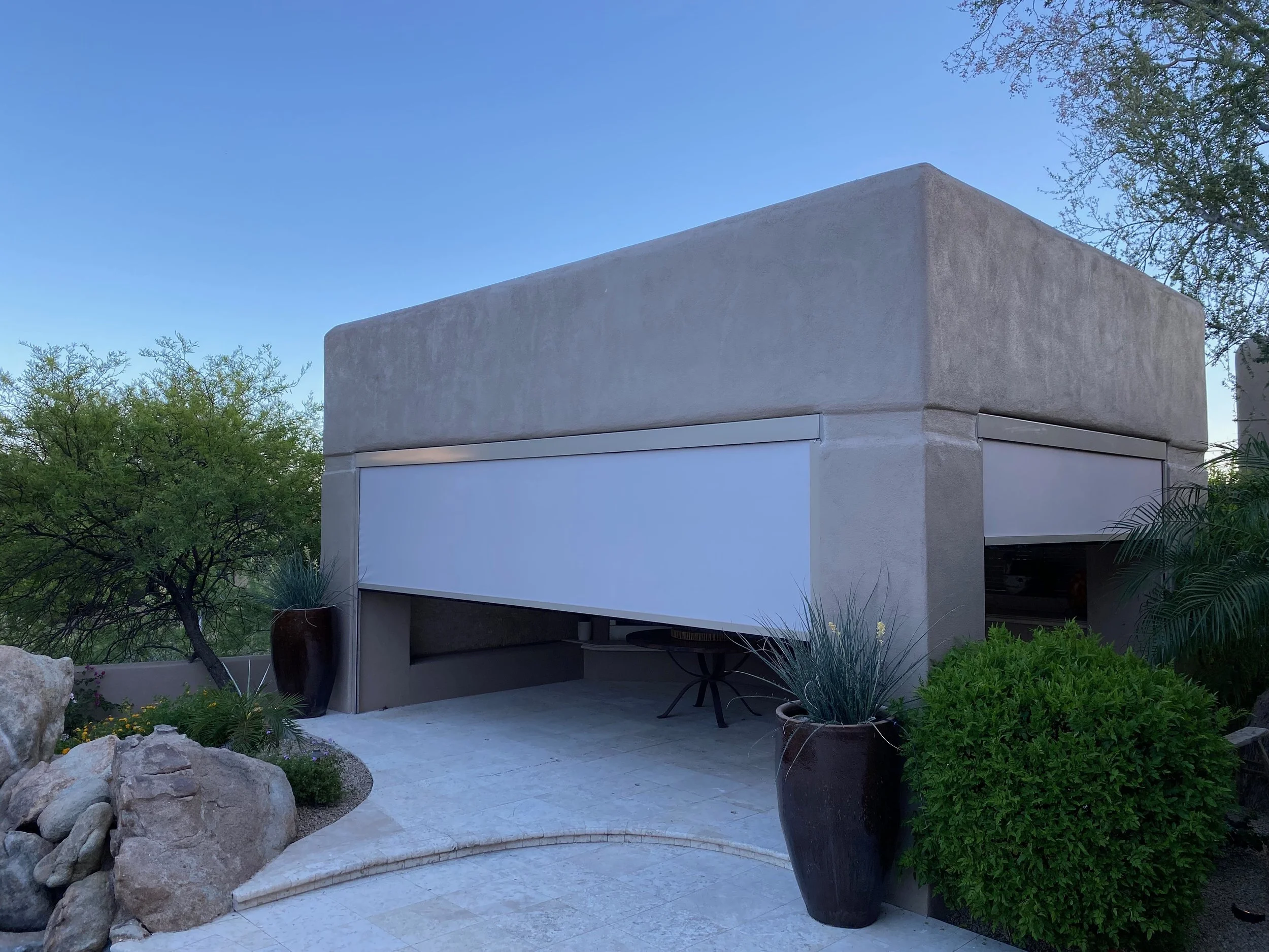 Modern house with a beige concrete exterior, large potted plants, and a minimalist patio under a clear blue sky.