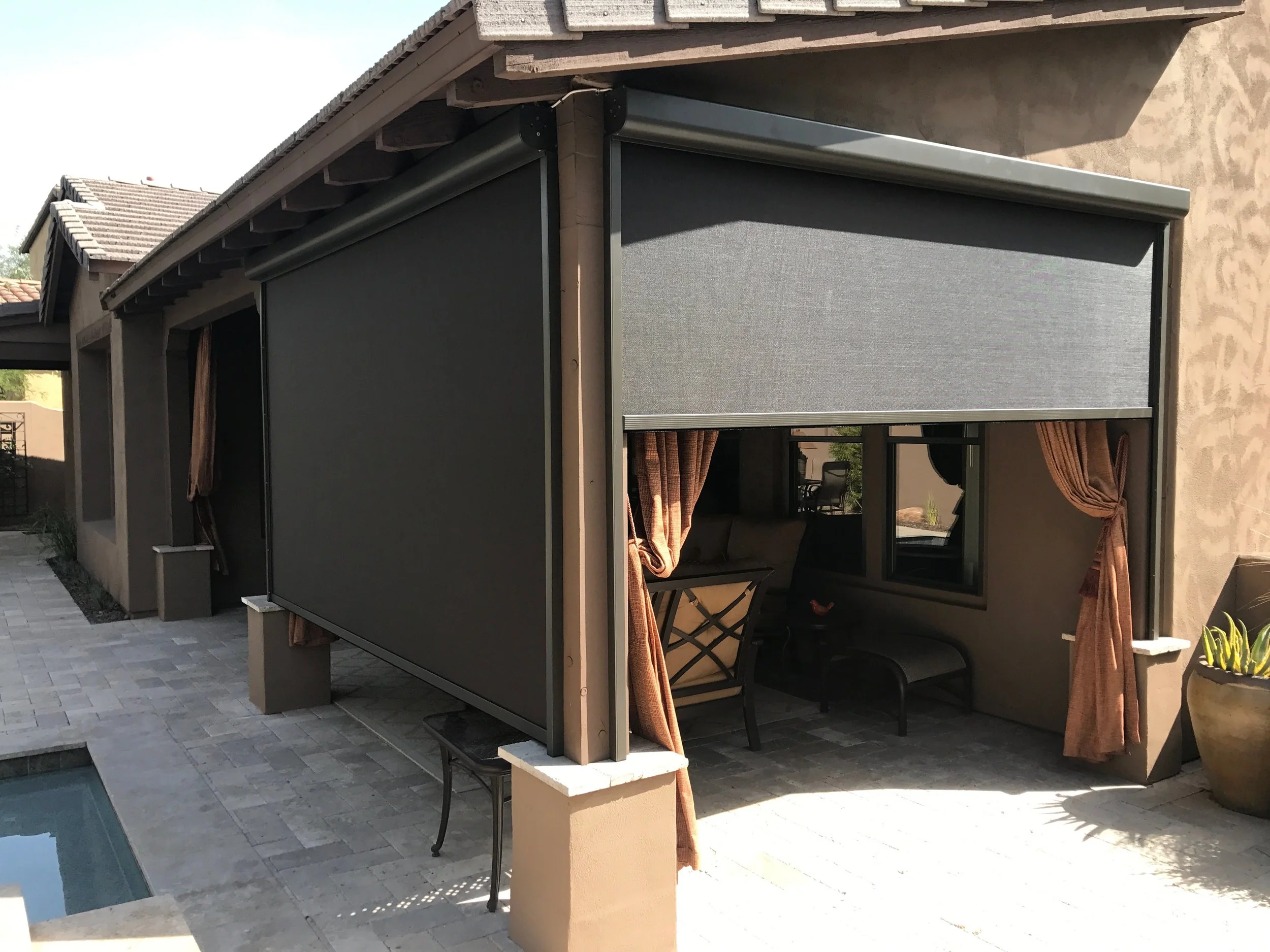 A backyard patio with a covered sitting area featuring black outdoor furniture, curtains, a small window, and a swimming pool to the left.