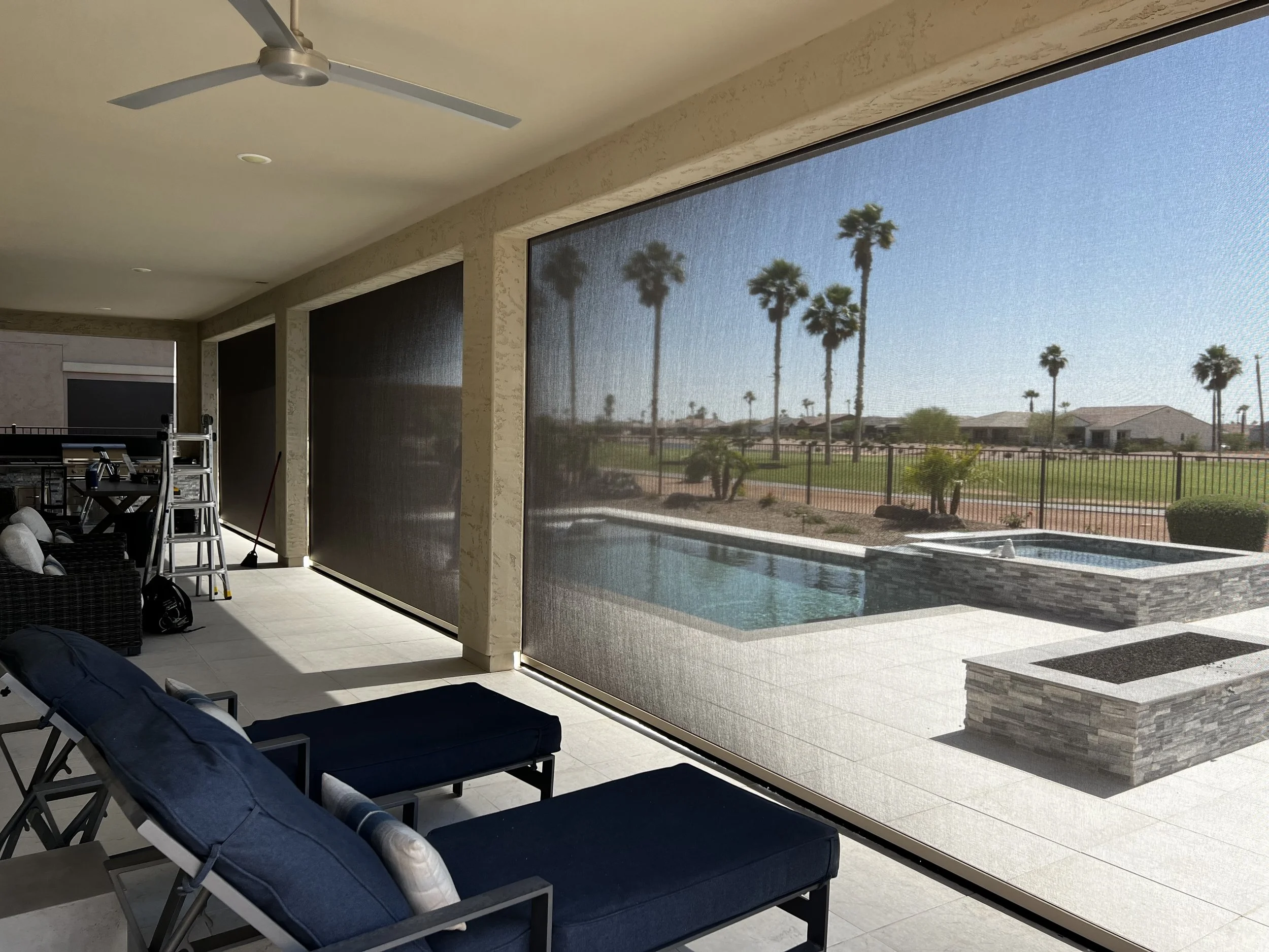 Covered patio with lounge chairs, a ceiling fan, and a view of a backyard pool and hot tub with palm trees beyond a fence in a sunny desert landscape.