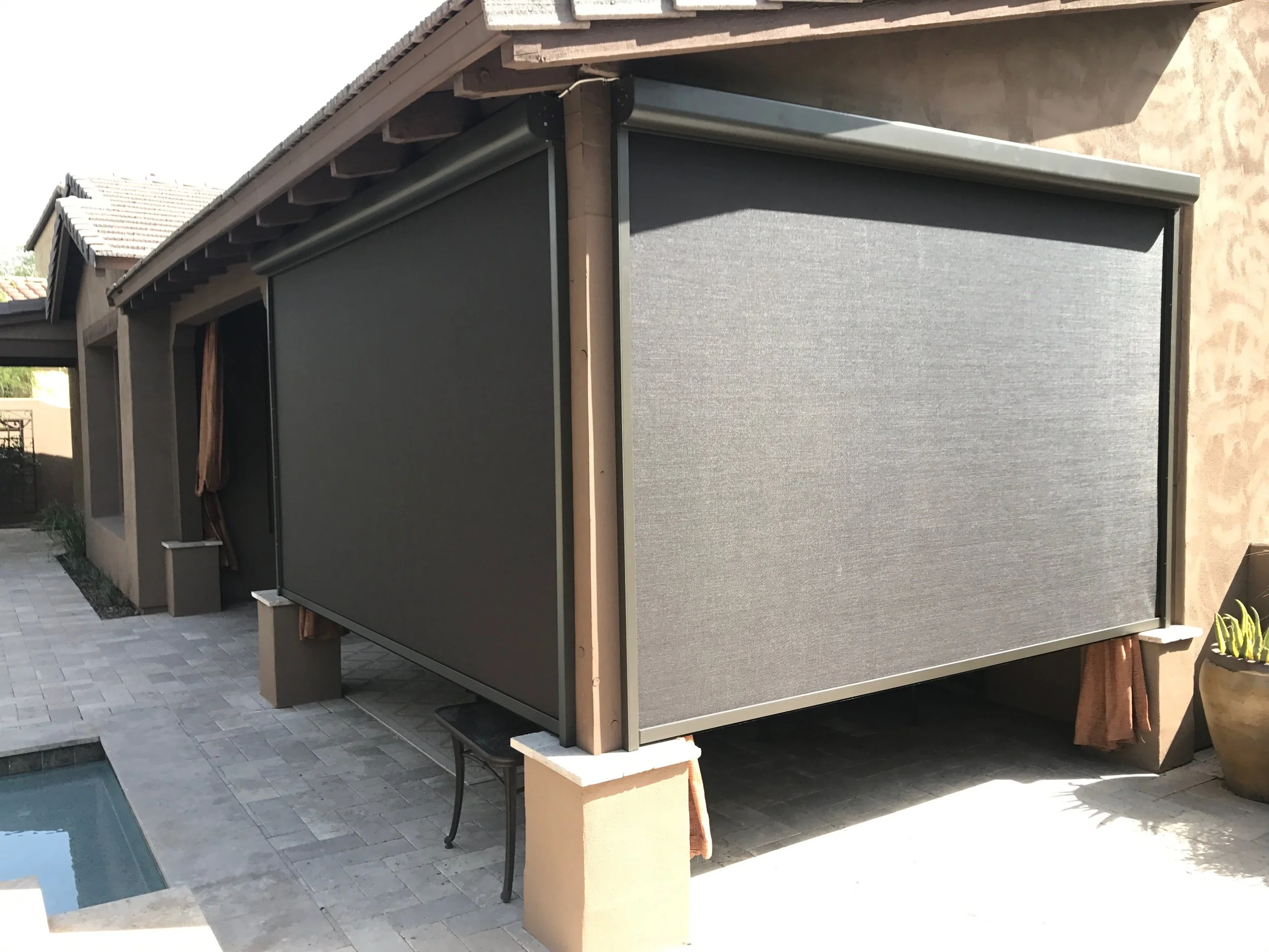 Back patio with gray retractable screen shades on the side of a house, small patio furniture, and a nearby potted plant.