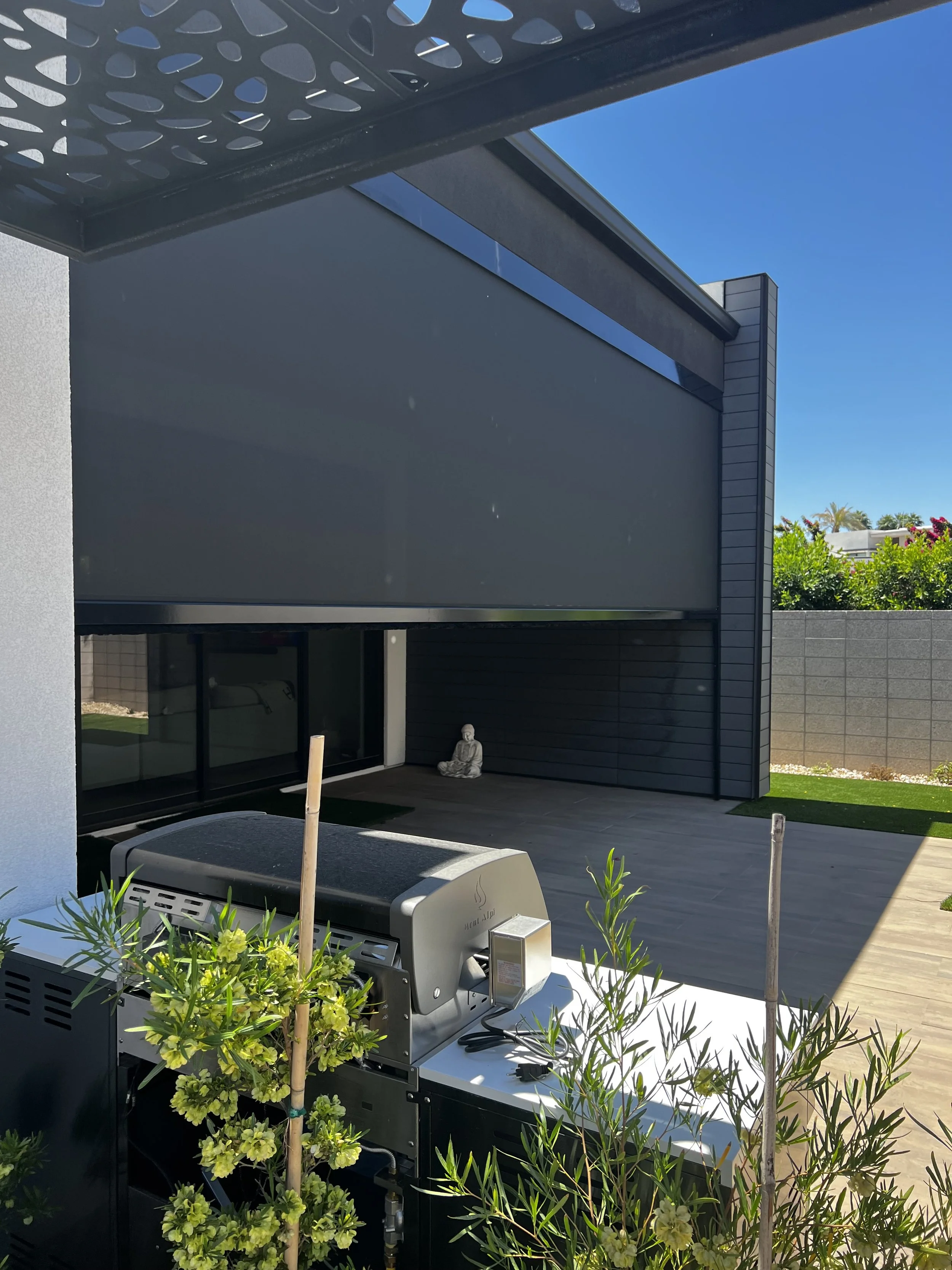 Outdoor backyard patio area with a grill, green plants, a small Buddha statue, and a modern building with black and gray exterior.