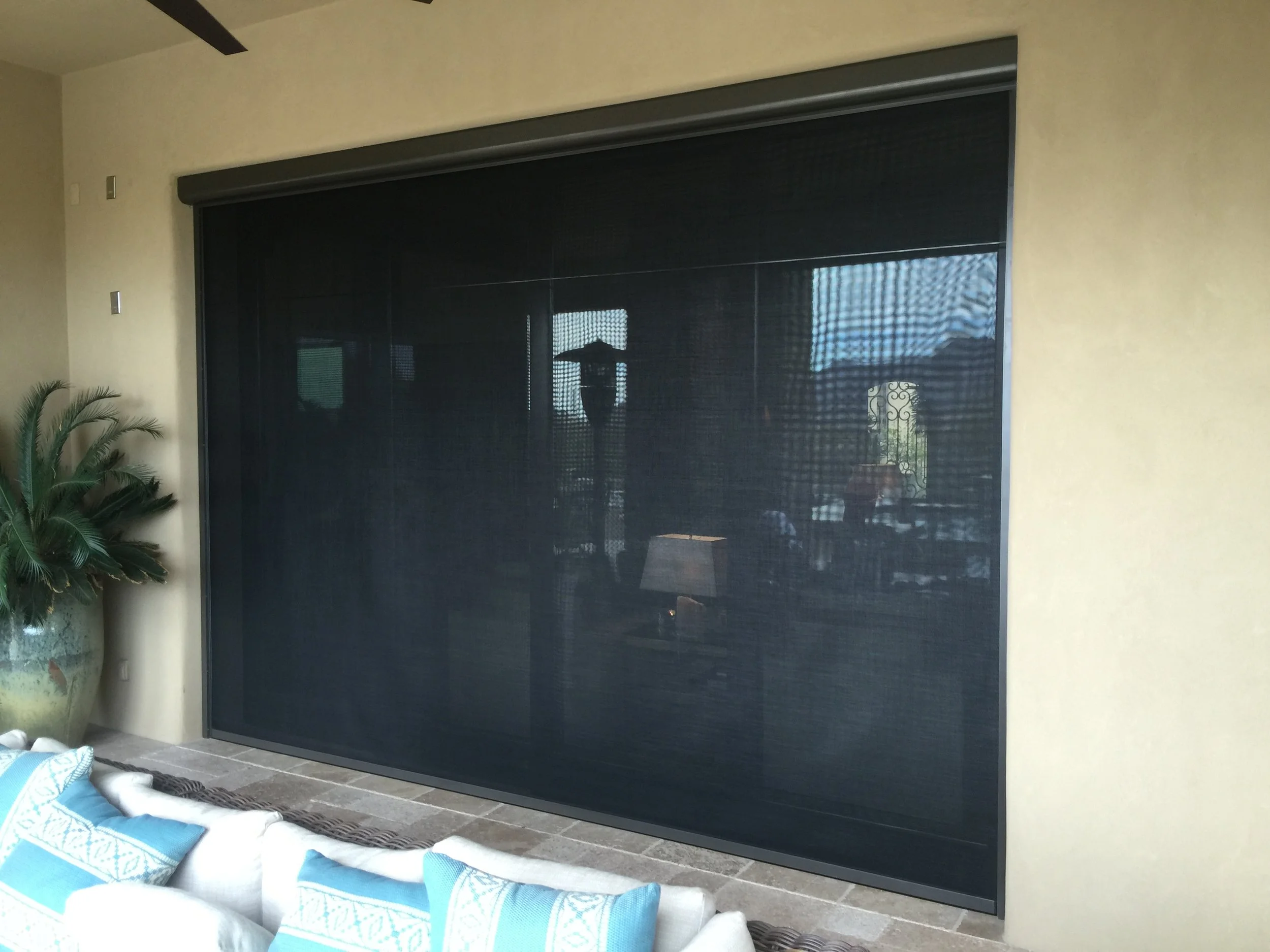 A large black retractable screen or door closed in front of a living room window, with a beige wall to the side, a potted plant on the left, and a sofa with blue and white pillows at the bottom of the image.