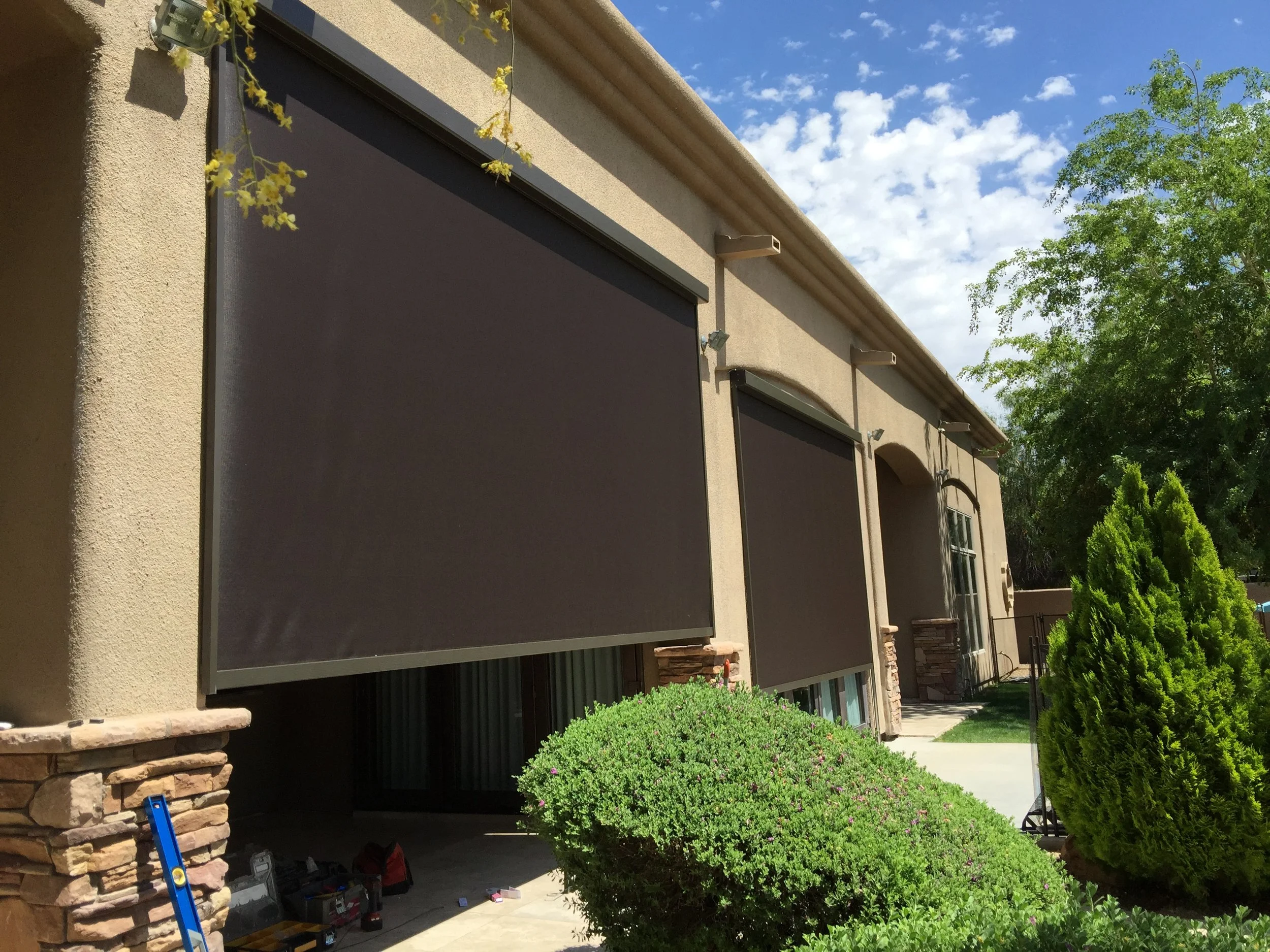 Exterior view of a house with two large black window shades rolled down, greenery, and a partly cloudy sky.