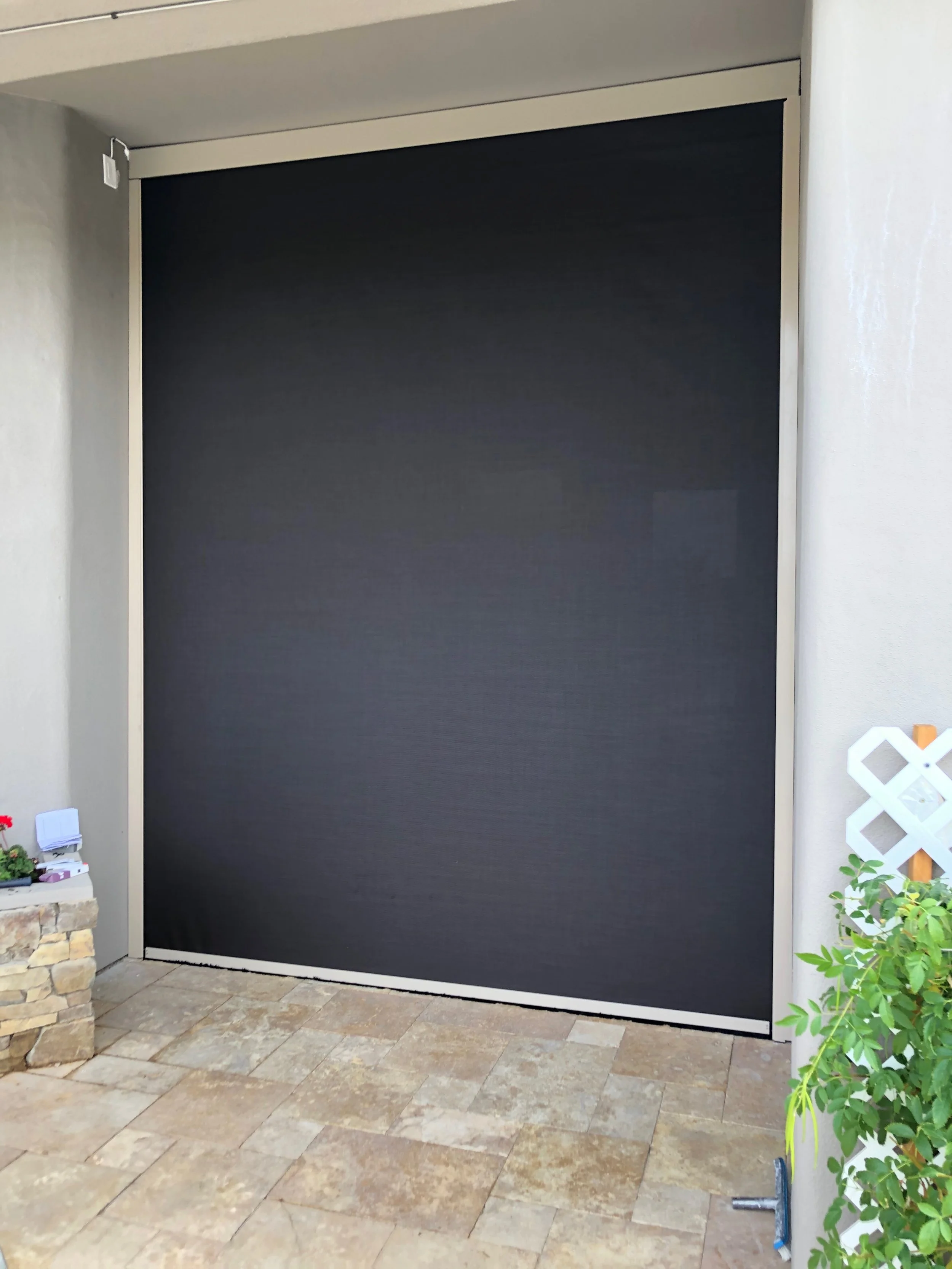 A large black garage door with a white frame installed on a beige exterior wall with tan stone flooring in front.